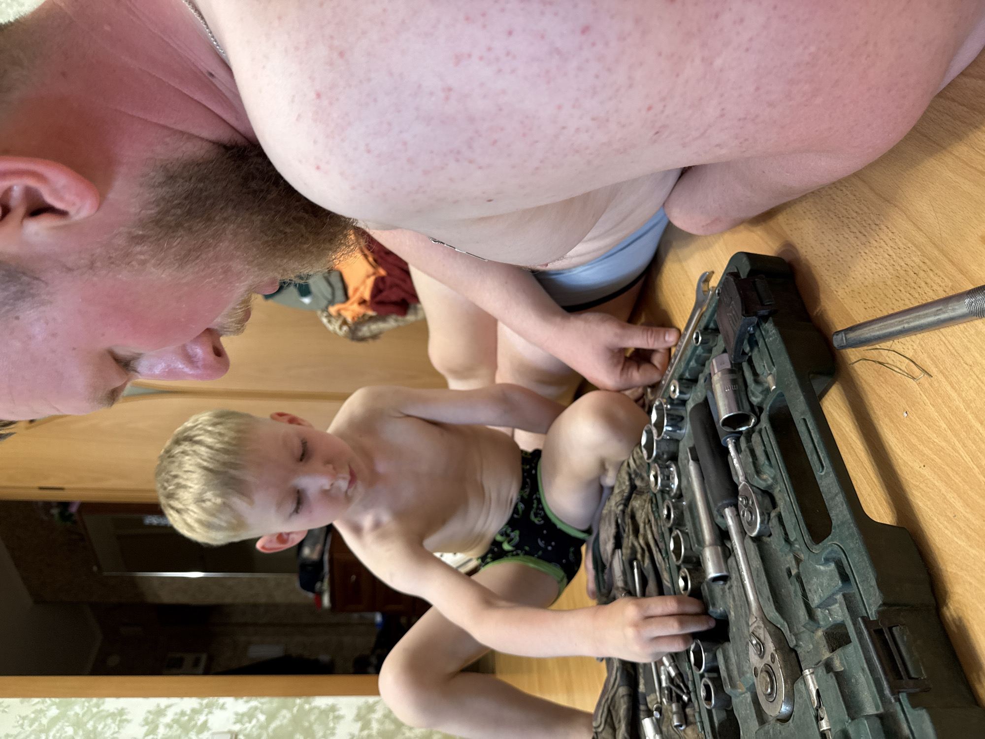 The image shows a man and a boy working on a tool kit. The man is on the left, seen from the side, with a beard and a partially visible body. The boy is in the center, also seen from the side, wearing dark shorts with green trim. He is bent over the tool kit, his hands engaged with the tools. The tool kit is dark green and filled with various sockets and ratchets, laid out on a light brown wooden surface. The scene appears to be indoors, with hints of a doorway and wooden furniture in the background. The lighting suggests it could be daytime. The overall atmosphere seems casual and focused on the task at hand.