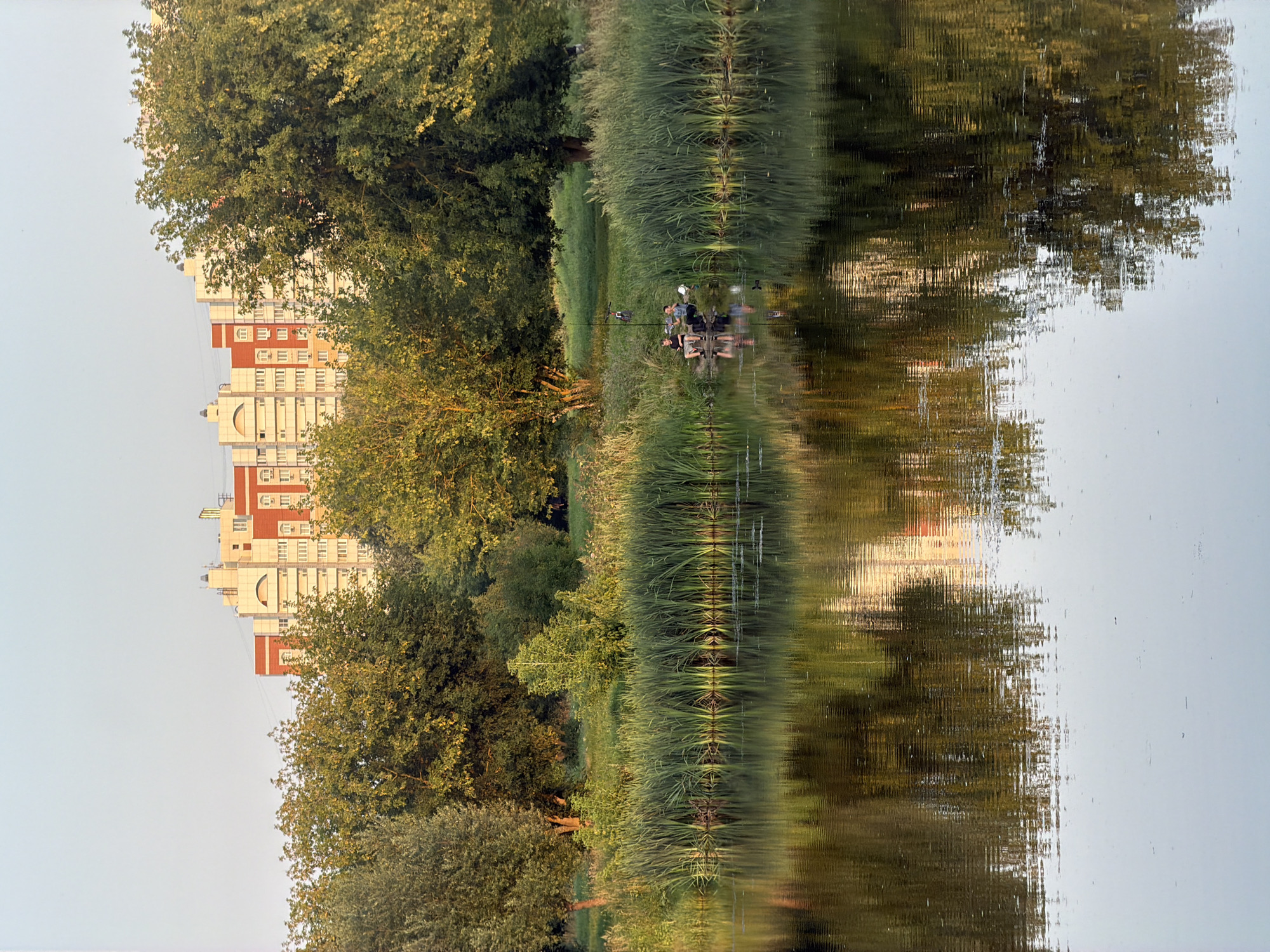 This is a photograph of a tranquil natural scene with an urban backdrop, likely taken in Kolomna, Russia. In the foreground, a calm body of water reflects the surrounding environment. Along the water's edge, tall, green reeds create a natural barrier. Beyond the reeds, a grassy area is dotted with trees, some of which are lush and green, while others have autumnal hues. In the middle ground, two individuals are seated by the water, appearing to be fishing. They are positioned near a fishing rod and some equipment. In the background, a large, multi-story apartment building with a distinctive red and white facade stands out against the clear, pale sky. The building's architecture is modern, with visible windows and decorative elements. The overall impression is one of a peaceful coexistence between nature and urban development.