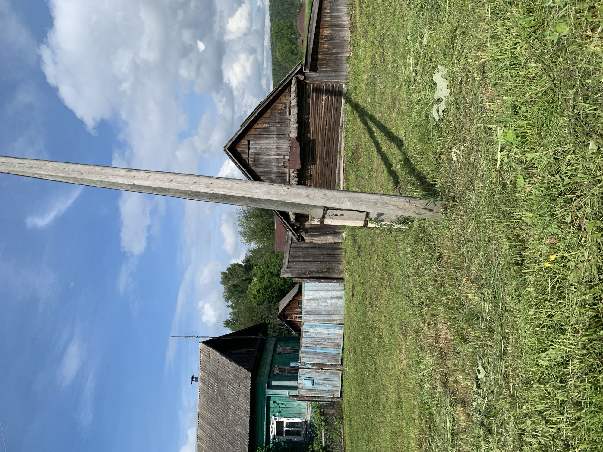This is a full-frame, eye-level, outdoor, daylight, medium shot of a rural scene. The background consists of a bright blue sky with scattered white clouds. In the mid-ground, there are several wooden structures, including a green wooden house with a grey roof on the left, and a rustic wooden barn-like building with a dark wood exterior and a grey roof on the right. A weathered blue and white wooden fence separates the green house from the rest of the yard. There are also several trees and vegetation in the background. In the foreground, a large, rough wooden utility pole stands vertically, casting a long shadow across the freshly cut green grass. The ground is covered in green grass, with some areas showing cut grass clippings. The scene appears to be a residential area in the countryside. The location is identified as Barsai, Russia.