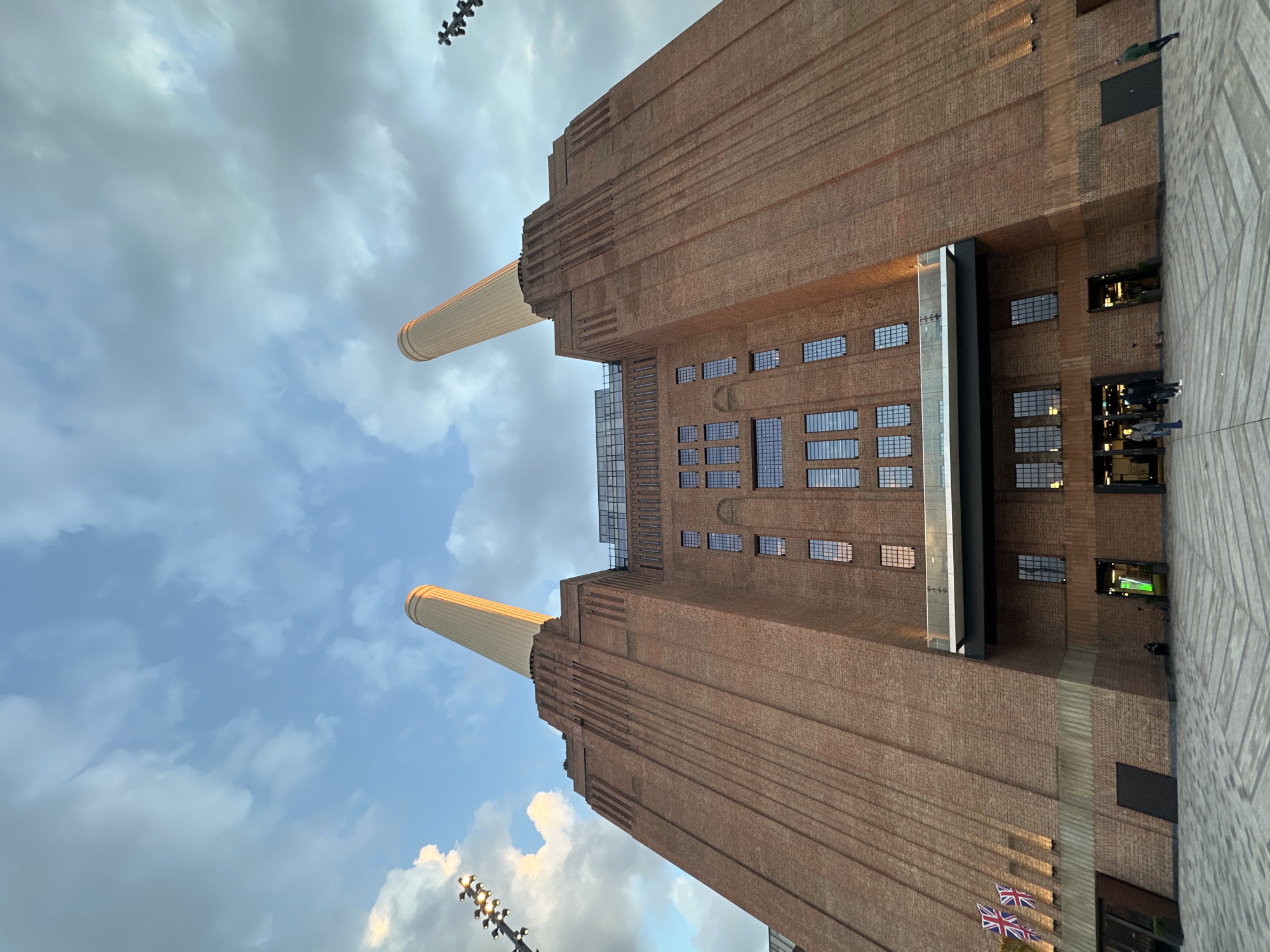 The image shows Battersea Power Station in London, United Kingdom, viewed from a low angle, with the blue sky and white clouds visible in the background. The red brick building with its four iconic chimneys dominates the frame. A portion of the pedestrian walkway to the right of the building has a few people walking. British flags are visible in the lower corner.