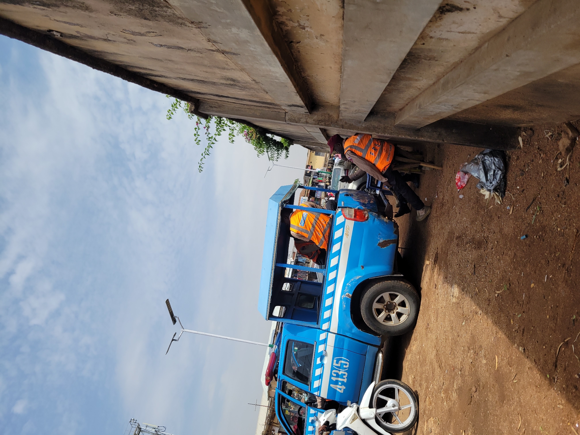 The image shows a blue traffic enforcement vehicle parked along the side of a road in Jos, Nigeria, next to a concrete structure, likely a bridge or overpass. Two individuals wearing orange safety vests are near the vehicle. One person is bending near the vehicle, while the other is standing in the back. The vehicle has a number "4-13(5)" painted on the side. A motorcycle is parked near the front of the vehicle. The environment consists of sky with clouds and a structure with steps and vegetation overhanging in the background. Debris like trash bags are seen on the ground.