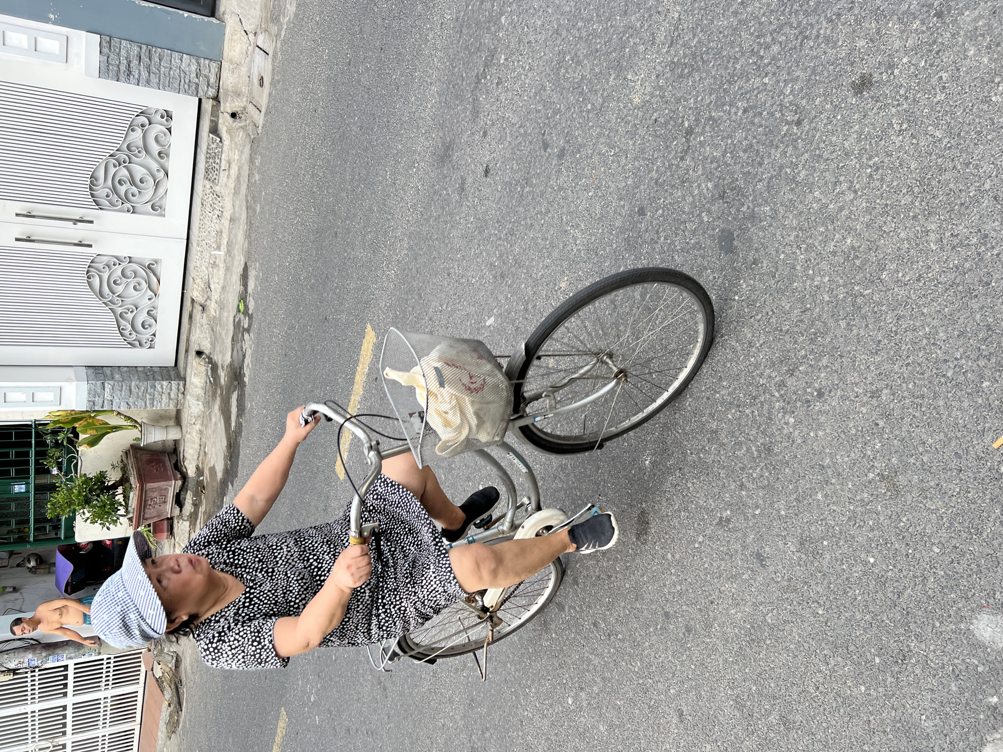 A woman is riding a bicycle down a street in Nha Trang, Vietnam. She is wearing a black and white polka dot dress, a checkered cap, and black sneakers. The bicycle has a basket on the front that contains a white bag. The street is paved with asphalt and has a yellow line on the left side. There are buildings and trees visible in the background. The image appears to be taken from a slightly elevated angle, looking down at the woman and her bicycle.
