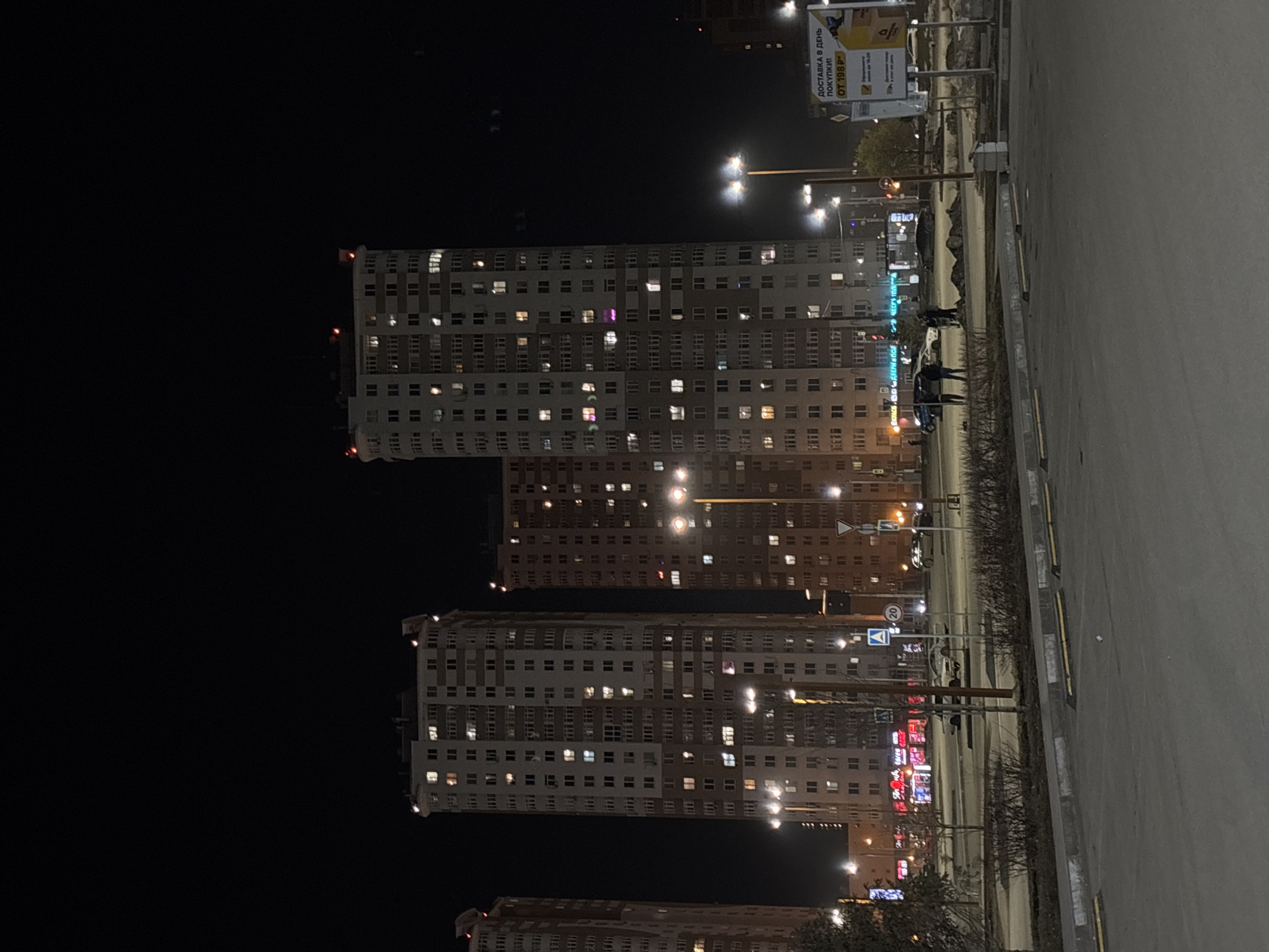 The image shows a night view of residential buildings and street lighting in Perm, Russia. The buildings are multi-story structures with numerous windows, many of which are illuminated. A parking lot is visible in the foreground, with cars parked along the street. Streetlights illuminate the road and parking area, and signage can be seen on a nearby building. Several people are standing on the street. The sky is dark.