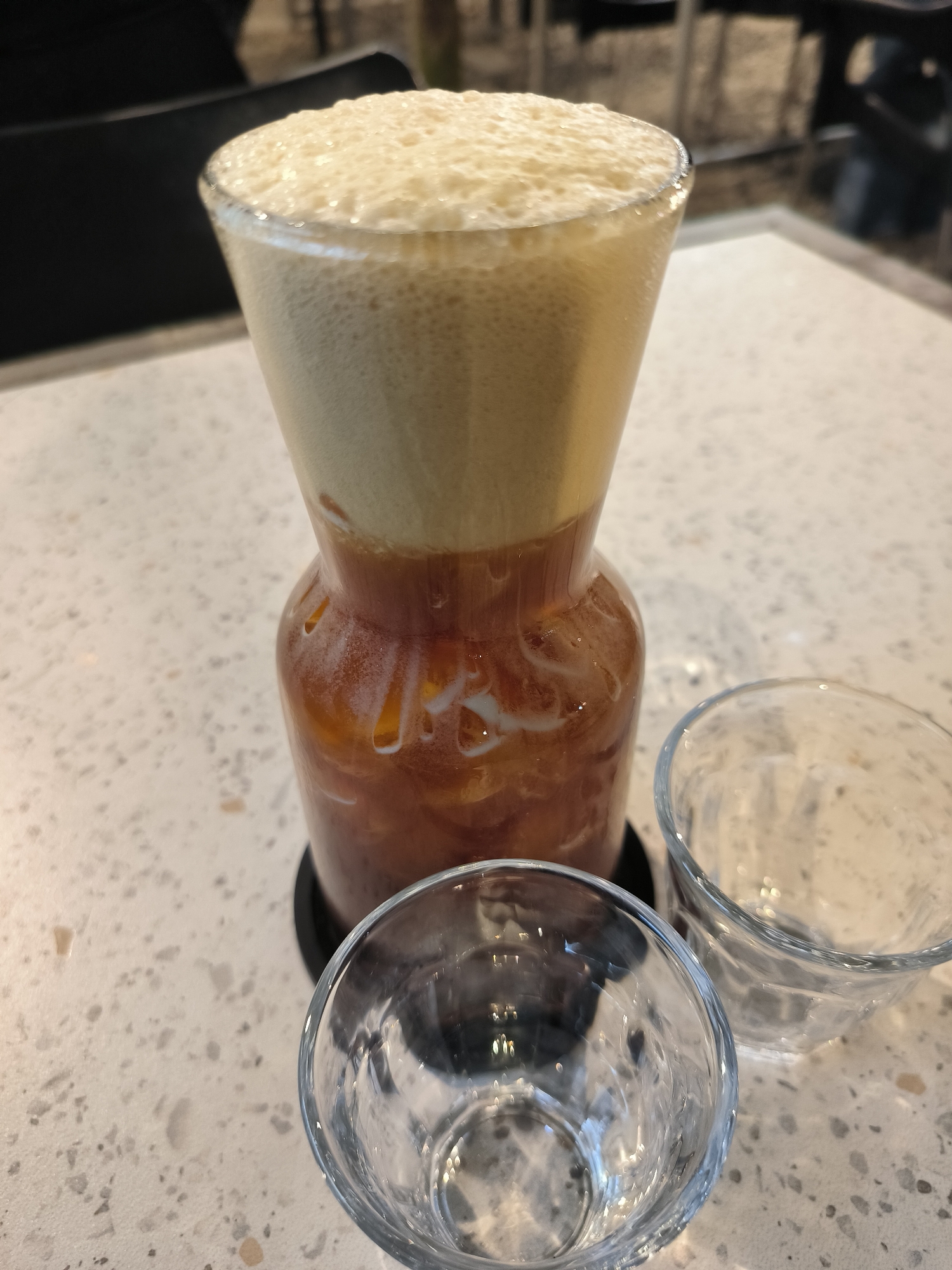 A pitcher of frothy iced tea sits on a speckled countertop. Two small, empty glasses are placed in front of the pitcher. The background is blurred, showing a dark chair and what appears to be a restaurant or cafe setting. The image is taken from a high angle, focusing on the drinks. No people are visible. The scene is likely indoors, given the lighting and furniture. The location of Lhokseumawe, Indonesia is not determinable from the visual information provided.