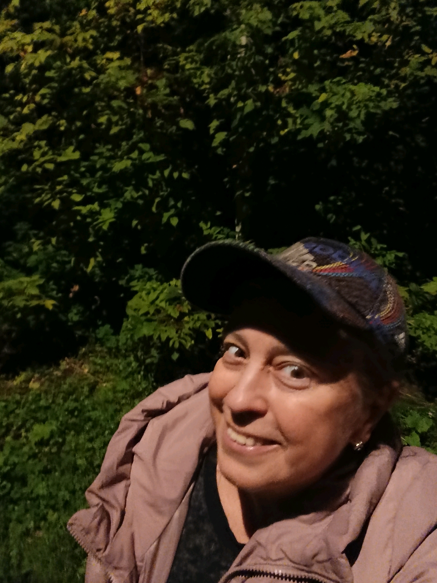 A woman wearing a baseball cap and a light-colored jacket is pictured outdoors at night, with a dense green foliage background, possibly in Verea, Russia. Her head is tilted, and she is smiling at the camera, with her eyes wide open. She is wearing a patterned baseball cap and a light pink or mauve jacket. The background consists of dark green leaves and foliage, illuminated by a light source that casts shadows. The lower part of the image shows the woman's upper body, including her jacket and a portion of her face and hat.