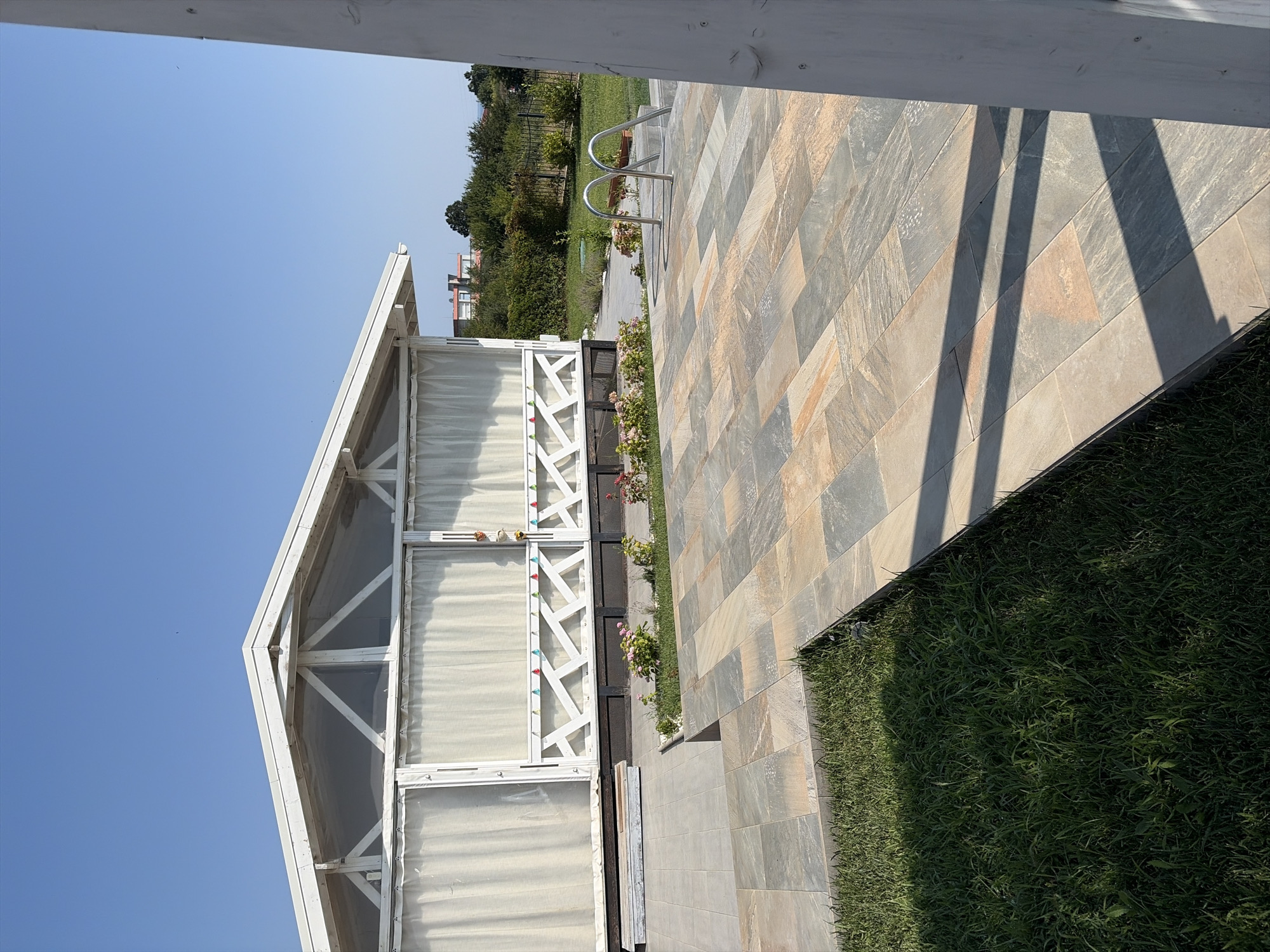 The image depicts an outdoor scene at a location in Velika, Bulgaria, likely a property with a swimming pool. In the foreground, a paved area made of rectangular stone tiles in shades of grey, beige, and orange stretches towards the right. To the left of the paved area is a strip of green grass.
Dominating the middle ground is a white, open-sided structure with a pitched roof, possibly a gazebo or covered seating area. This structure has white curtains drawn across its openings. A decorative lattice work runs along the bottom of the structure's facade, adorned with small, colorful triangular flags. There are flower beds with pink and red flowers planted in front of the structure.
To the right of the paved area, a portion of a swimming pool is visible. It has a set of silver-colored metal handrails leading down into the water, which is not fully visible. The pool appears to be above ground, with a concrete or tiled surround.
In the background, beyond the structures and pool, is a green lawn bordered by a black metal fence. Further in the distance, trees and some buildings are visible, with a clear blue sky overhead. The lighting suggests it is daytime, with shadows cast by the structures onto the paved area. There are no people visible in the image.