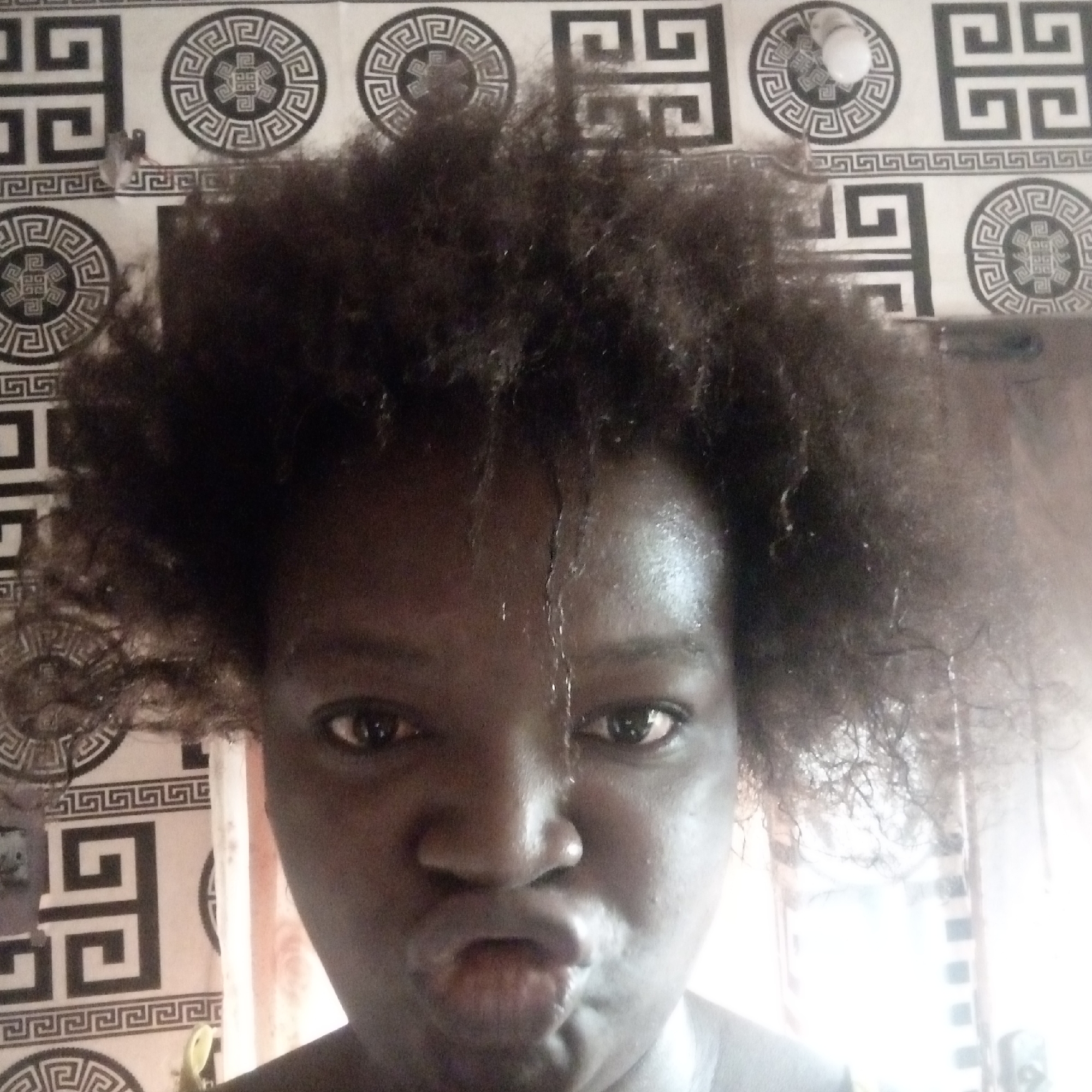 A medium-close portrait of a Black female with dark eyes and short, curly dark hair, taken at an upward angle. She is pouting her lips slightly. A vertical streak of moisture or sweat is visible on her forehead. The background consists of a light-colored wall covered in a repetitive pattern of geometric shapes in black and dark grey, including circles and rectangles. To the left of the subject, a portion of a white curtain is visible. The photograph seems to have been taken indoors, with ambient light. The location of Onitsha, Nigeria is not evident from the image's visual information.