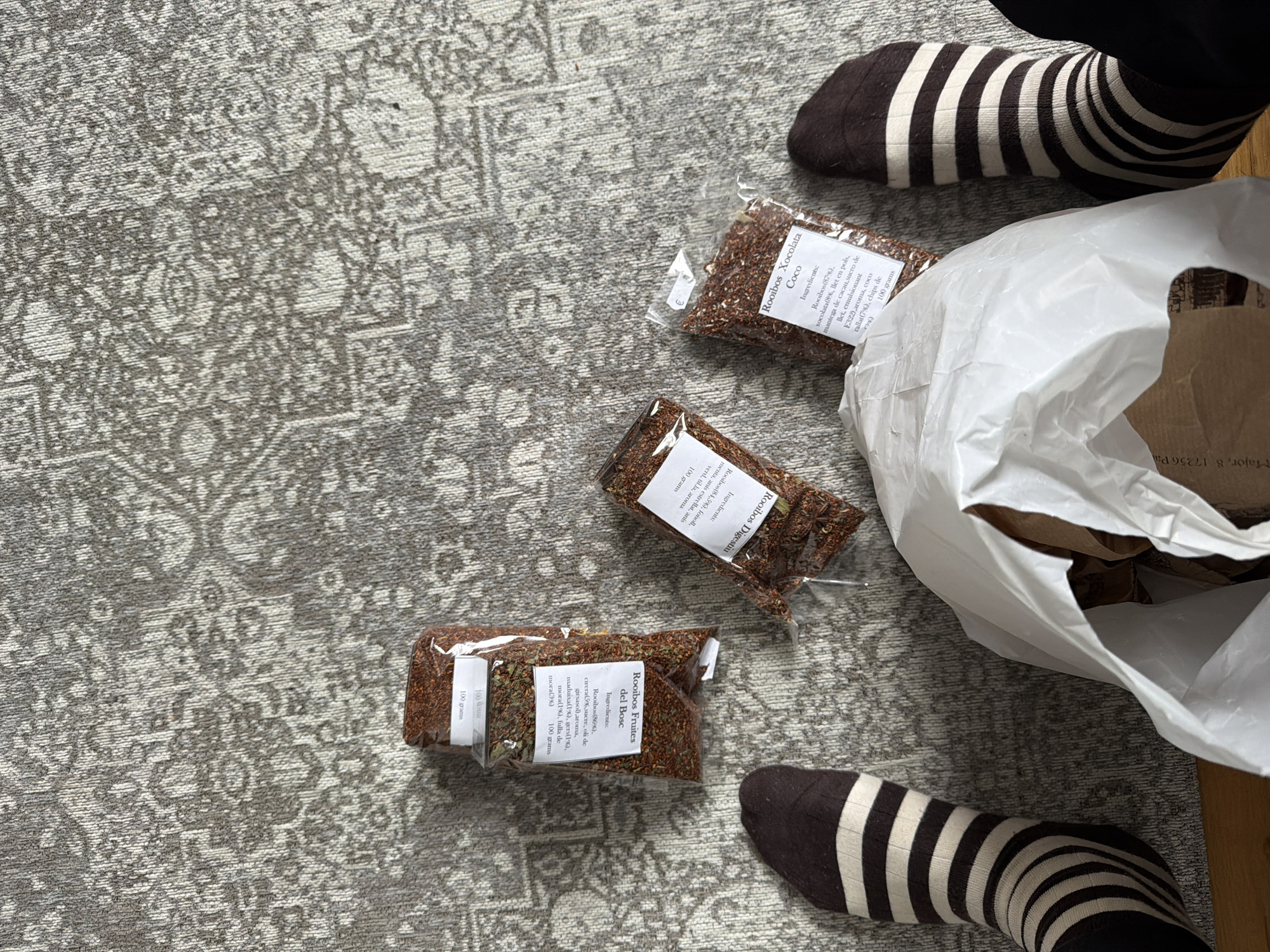 The image shows a gray patterned rug with three sealed packages of rooibos tea. A white plastic bag partially hides one of the packages. Each package has a label with the name of the tea and the ingredients written in both Catalan and English, and the weight is 100 grams. The three varieties of tea are "Rooibos Xocolata Coco", "Rooibos Digestiu", and "Rooibos Fruites del Bosc". The photo also shows a person's feet wearing black socks with white stripes, the left foot is to the bottom-left of the packages and the right foot is to the top-right of the packages. The address "Major, 8 17256 Pa" is partially visible in the plastic bag.