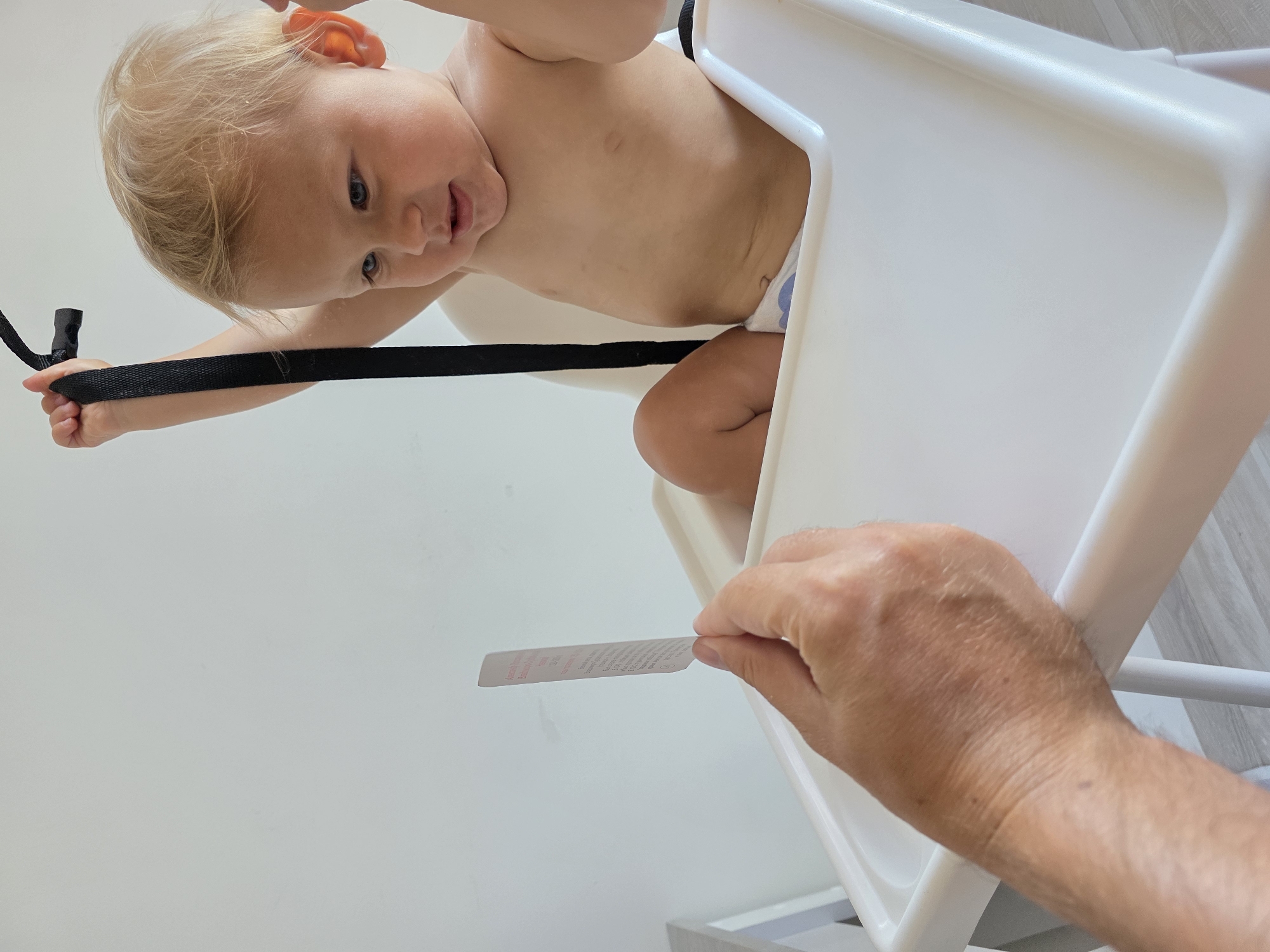 The image shows a close-up, tilted shot of a baby sitting in a white high chair. The baby has blonde hair and blue eyes and appears to be looking towards the camera with a slight smile. The baby is wearing a diaper and has a black strap from the high chair in its left hand. A person's hand is visible in the lower right corner, holding a small white tag with some red text on it. The background is a plain white wall. The image appears to be taken indoors during the daytime. The overall mood is casual and domestic.