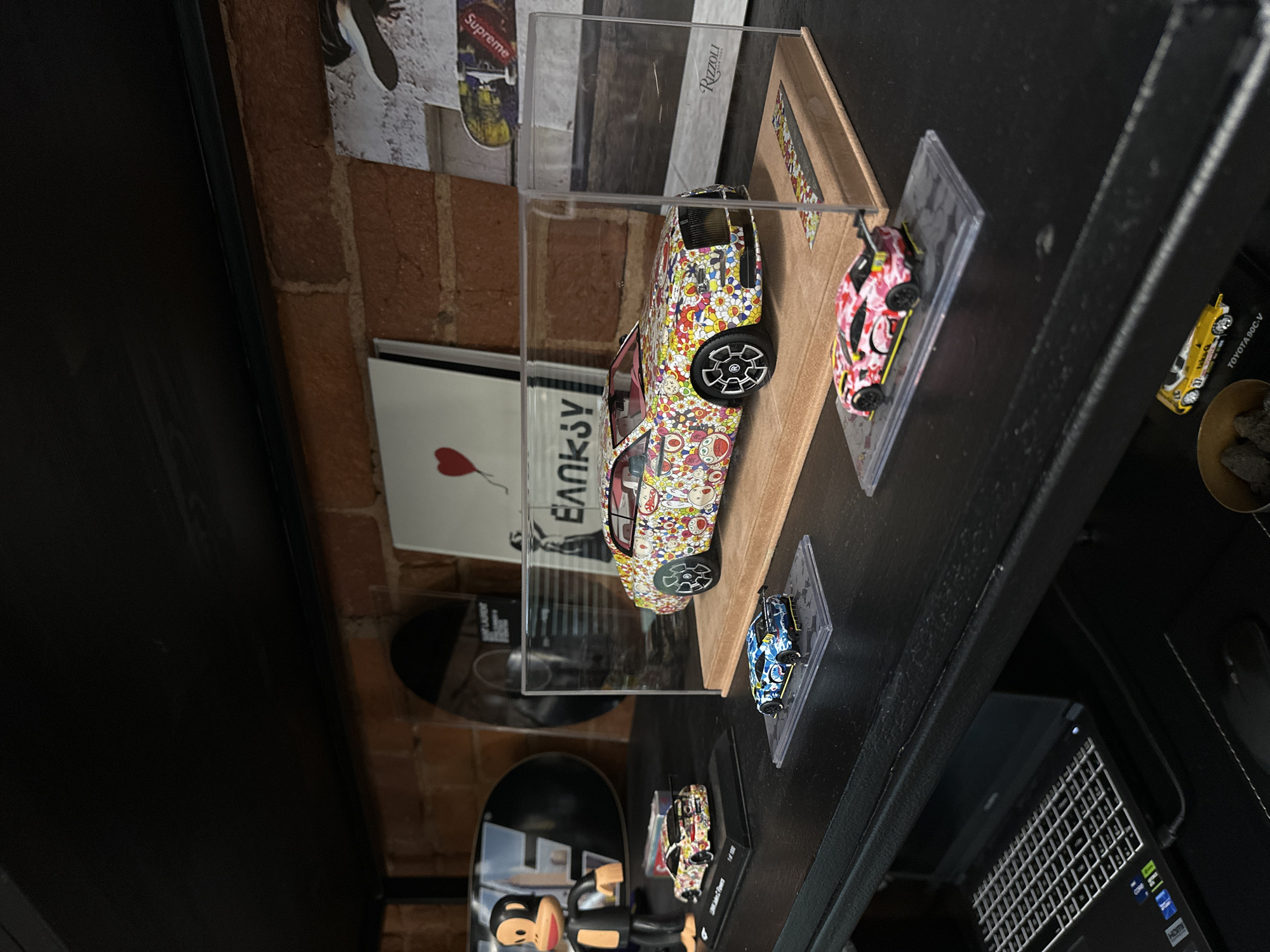 A still, close-up shot captures a display of model cars, seemingly housed in a room with exposed brickwork, possibly in Moscow, Russia. Several model cars, encased in clear plastic display cases, are arranged along a dark shelf or ledge. One larger, more prominent model car is enclosed within a larger acrylic box, elevated on a wooden base. Adjacent to the cars, there are framed artworks, including one featuring the word "Banksy" with a heart motif. In the lower right of the frame, part of a laptop keyboard is visible. A Supreme sticker is present on the wall above. The lighting is slightly dim, accentuating the objects on display against the backdrop of the room's architecture.