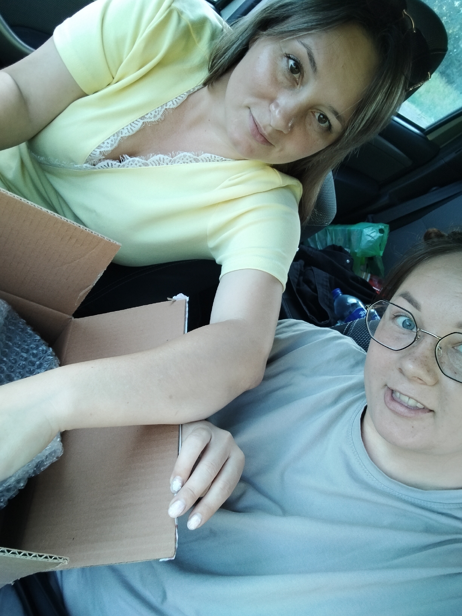 Two women are inside a car, with one woman in the foreground and the other in the background. The woman in the foreground is wearing a light blue t-shirt and glasses, and is looking at the camera with her mouth open. The woman in the background is wearing a yellow top with white lace trim and is also looking at the camera. Both women appear to be smiling.

In front of the woman in the foreground is a cardboard box. Part of the box is open, and some bubble wrap is visible inside. The car's interior is visible, including seats and some bags in the background. The lighting suggests it is daytime. The overall mood appears casual and friendly. No visible text is present in the image. The location context is Lunino, Russia.