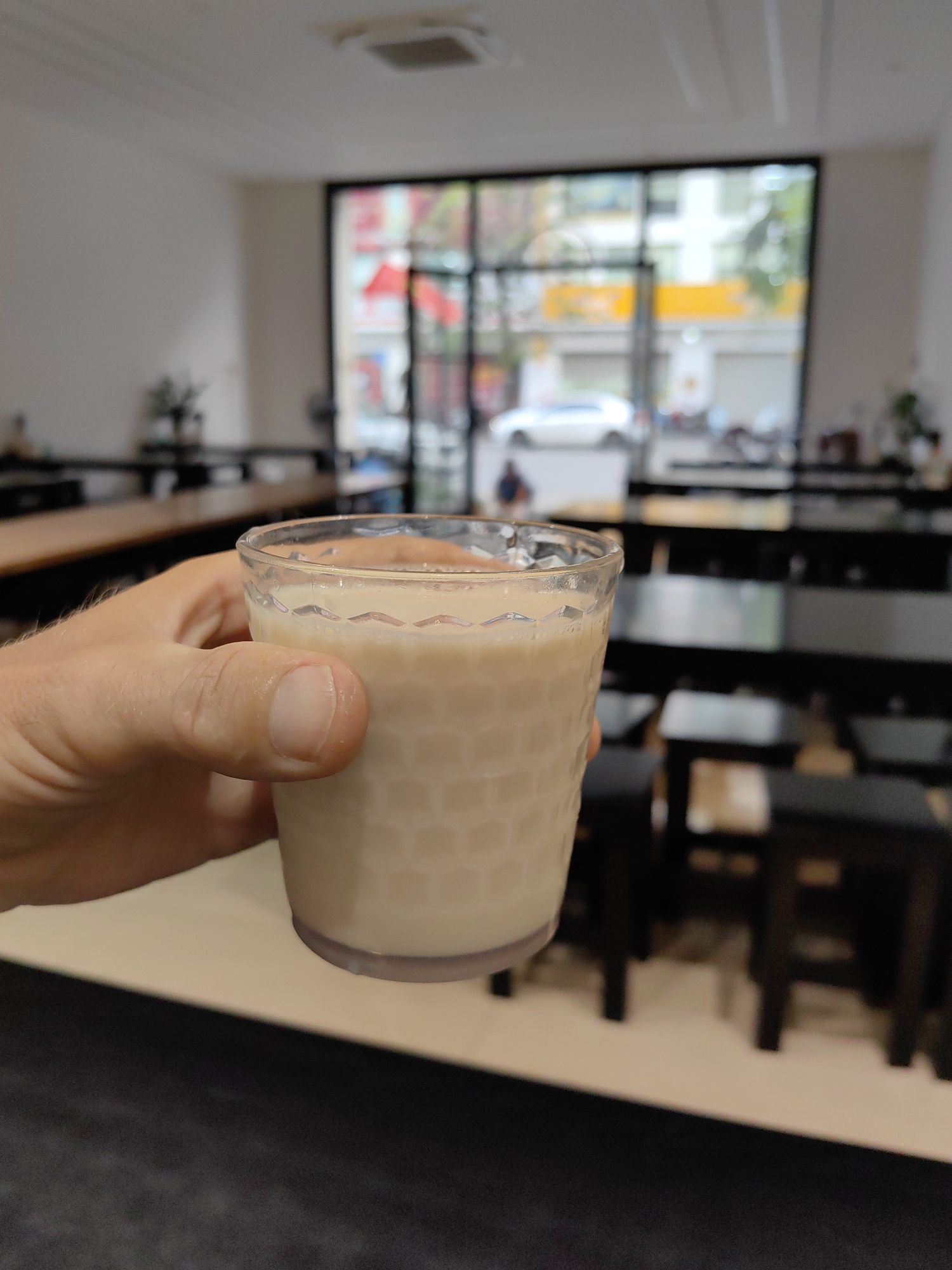 A first-person view captures a hand holding a glass of light brown liquid. The glass has a patterned surface and is almost full. The background is a restaurant interior with long wooden tables and black stools. A large window reveals an exterior scene, including a white car, motorbikes, and buildings, suggesting a street in Hong Gai Ward, Vietnam.