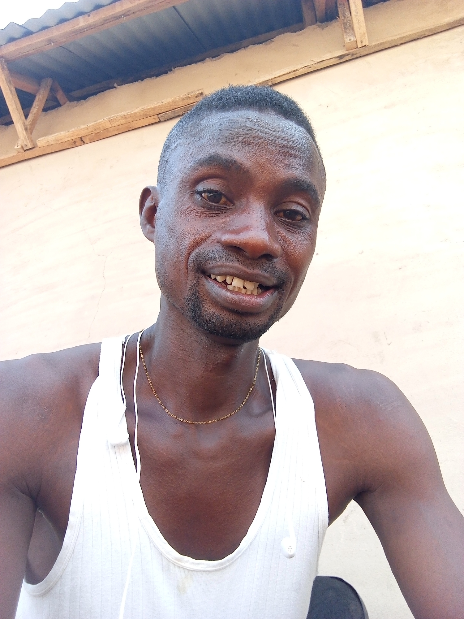 This media file is a close-up portrait of an adult male in Adonkolo, Nigeria.
**Subjects:** The central subject is a man with a dark complexion, short dark hair, and a short beard or stubble. He is wearing a white ribbed tank top (singlet) and a thin gold-colored chain around his neck. White wired earbuds are visible around his neck and shoulders. In the background, there's a light-colored wall, and above it, a roof structure with dark corrugated metal sheets and wooden support beams is partially visible.
**Activity or event taking place:** The man is posing for the camera, looking directly at the viewer and smiling broadly.
**Notable details:** The image is brightly lit by natural light, suggesting it was taken during the daytime under clear weather conditions. The man's wide smile and engaged eyes convey a sense of happiness and friendliness. The visible architectural elements (corrugated roof, wooden beams, plain wall) suggest a casual outdoor setting, possibly in a residential or undeveloped area. There is no visible text in the image.