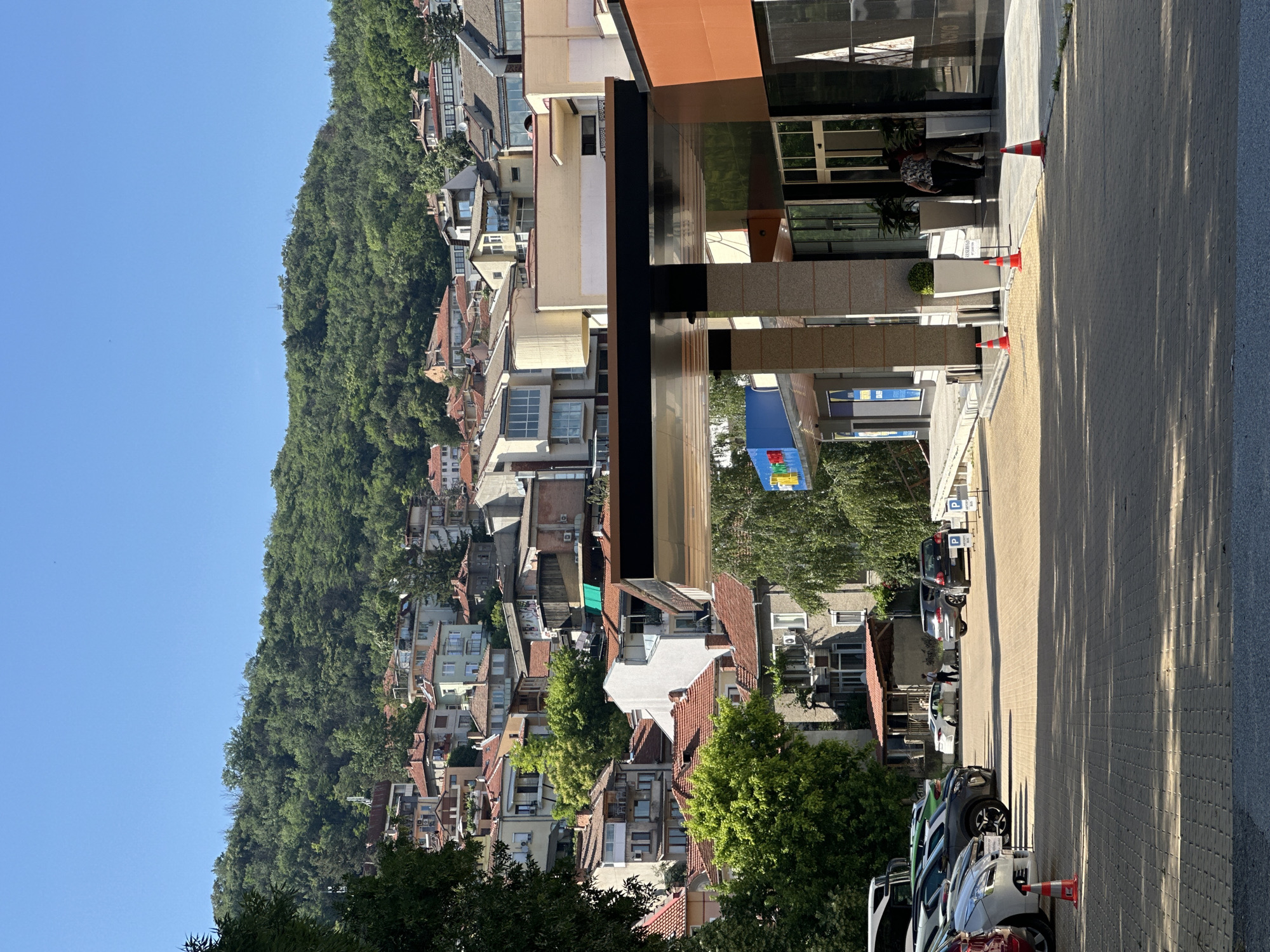 The image depicts a daytime scene in the city of Veliko Tarnovo, Bulgaria, characterized by a steep, tree-covered hillside populated with numerous residential buildings under a clear blue sky. In the foreground, a paved surface, likely a parking lot, features the partial view of several parked vehicles, including white, red, and various shades of grey cars. An orange traffic cone is visible in the far left foreground.
In the midground, on the right, stands a modern commercial building with a distinctive brown and orange facade and a large, dark, overhanging canopy supported by pillars. Its reflective underside is visible. Two individuals, one wearing a patterned shirt and dark pants, are captured entering or exiting the building's glass doors. To the left of this structure, another commercial building with a prominent blue sign is visible, accessible via a paved ramp. Further left, in the midground, two people stand near a white car next to an older, smaller building with a red-tiled roof and a wooden porch. Three additional orange traffic cones are placed near the entrance of the modern building on the right. Shadows are cast across the paved ground by overhead trees and structures, indicating strong sunlight. Two white "P" parking signs, one indicating "BUS," are visible on the right side of the parking area.
The background is dominated by a densely built hillside, where numerous houses and multi-story apartment buildings ascend the slope. These structures exhibit varied architectural styles, predominantly featuring reddish-brown tiled roofs, and facades in white, beige, light green, and grey tones. The upper reaches of the hillside are covered in dense, verdant forest. A faint, slender tower-like structure is discernible on the distant hilltop against the clear sky.