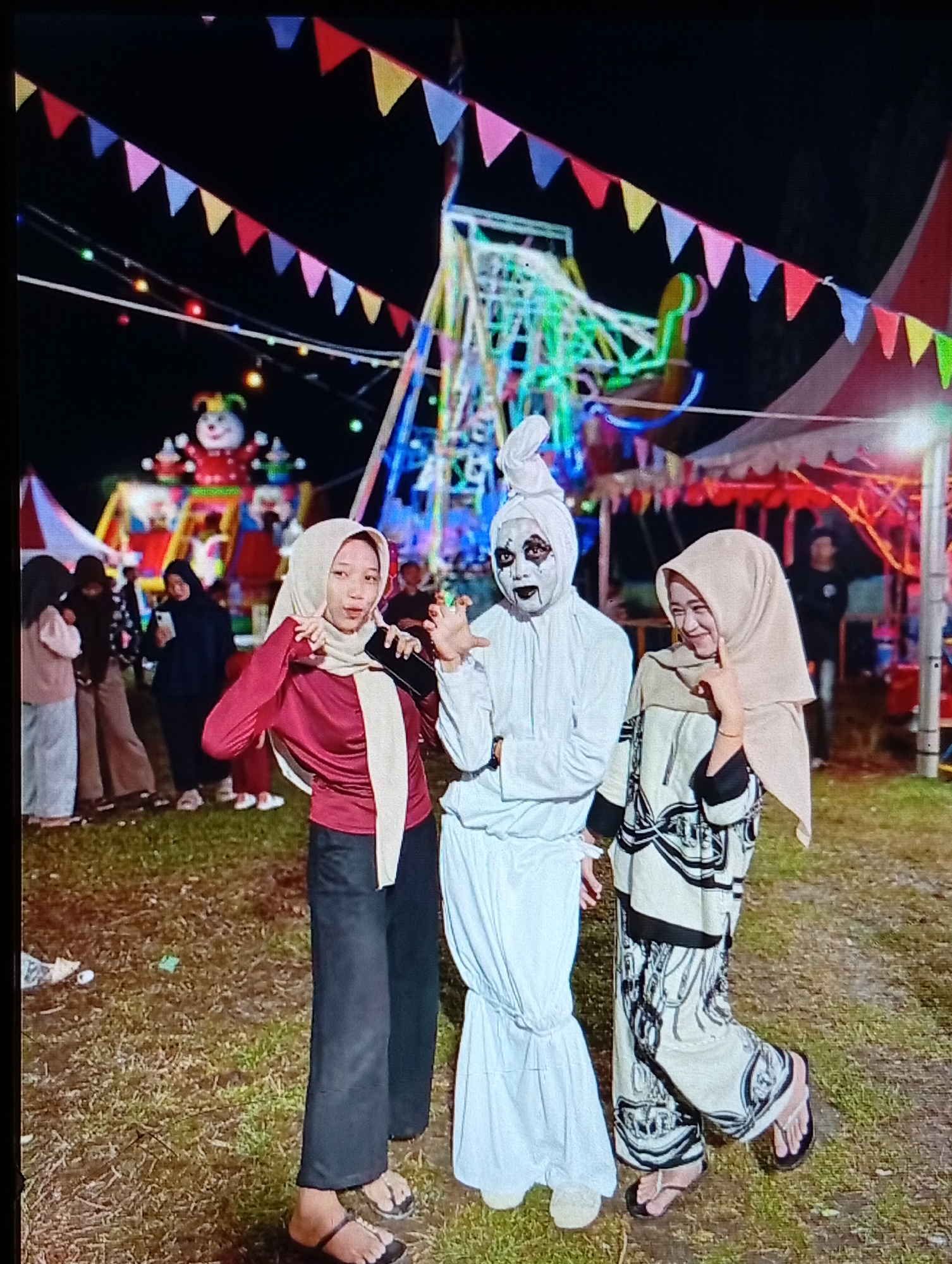 The image captures a nighttime fairground scene in Lhokseumawe, Indonesia. Three individuals are positioned centrally in the foreground, posing on an uneven ground composed of dirt and dry grass.
The individual on the left is a young female wearing a maroon long-sleeved top, dark wide-leg pants, and a light beige hijab. She is wearing dark flip-flops. Her left hand holds a black smartphone, and her right hand is positioned near her face. She is smiling subtly and looking towards the camera.
The central figure is an individual dressed in an all-white costume, resembling a ghost or mummy. The face is painted white with black markings around the eyes and mouth. A white fabric is wrapped on the head, forming a peak. Both hands are raised slightly, open-palmed, towards the camera. White footwear is also visible.
The individual on the right is a young female wearing a light beige hijab, a long-sleeved tunic with intricate black and white abstract patterns, and dark wide-leg pants. She is wearing dark flip-flops. She is smiling and looking towards the camera, with her right hand near her chin.
In the background, multiple other individuals are visible, some blurred, indicating a crowd at the event. Overhead, colorful triangular pennants (bunting) are strung across the frame. Various string lights with small, colorful bulbs are also present. Dominating the background on the right is a tall structure resembling a Ferris wheel, illuminated with vibrant blue and green lights. To the left, another illuminated structure features a large clown figure with a red body and multi-colored details. Portions of several tents, including one with red and white stripes on the far right, are visible. The scene is brightly lit by artificial light sources from the rides and decorations, indicating it is night.