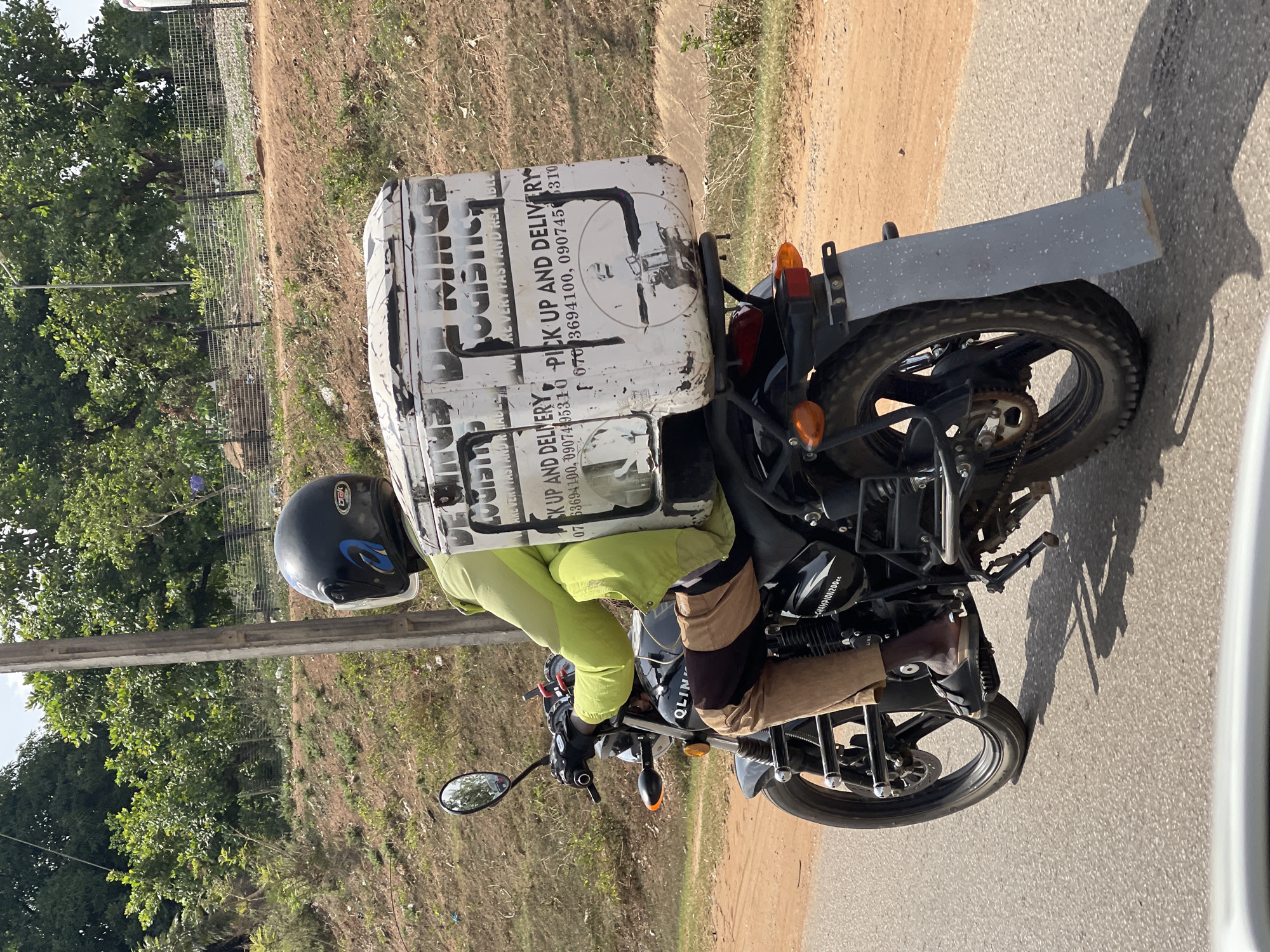 An outdoor, eye-level shot captures a person on a motorcycle, riding along a paved road next to a dirt shoulder, likely in Galadimawa Village, Nigeria. The scene is brightly lit by natural sunlight, casting visible shadows, indicating daytime with clear weather.
The primary subject is a person wearing a black helmet with blue accents, a lime green long-sleeved top, and multi-toned brown pants, riding a black motorcycle. A large, white delivery box is secured to the back of the motorcycle, featuring black stenciled text. The person is holding the handlebars, and their right leg is visible, with a foot resting on the motorcycle's footpeg. The motorcycle has a black body, silver accents, and an exposed chain drive visible on the rear wheel. A makeshift, grey rectangular mudguard is attached above the rear wheel.
The delivery box prominently displays text such as "LOGISTICS," "PICK UP AND DELIVERY," and phone numbers: "070 3694100, 09074-95310" and "070 3694100, 0907450310". Other phrases like "ARE VERY FAST AND RELIABLE" are also partially visible. On the motorcycle's engine casing, the text "CHAMPION200cc" and "QLIN" can be seen.
The background shows a simple roadside environment. To the left of the motorcycle is a dirt shoulder with some sparse vegetation and a metal mesh fence running parallel to the road. Beyond the fence, a dense cluster of green trees is visible. The road itself is asphalt, and the adjacent shoulder is dry, reddish-brown earth.