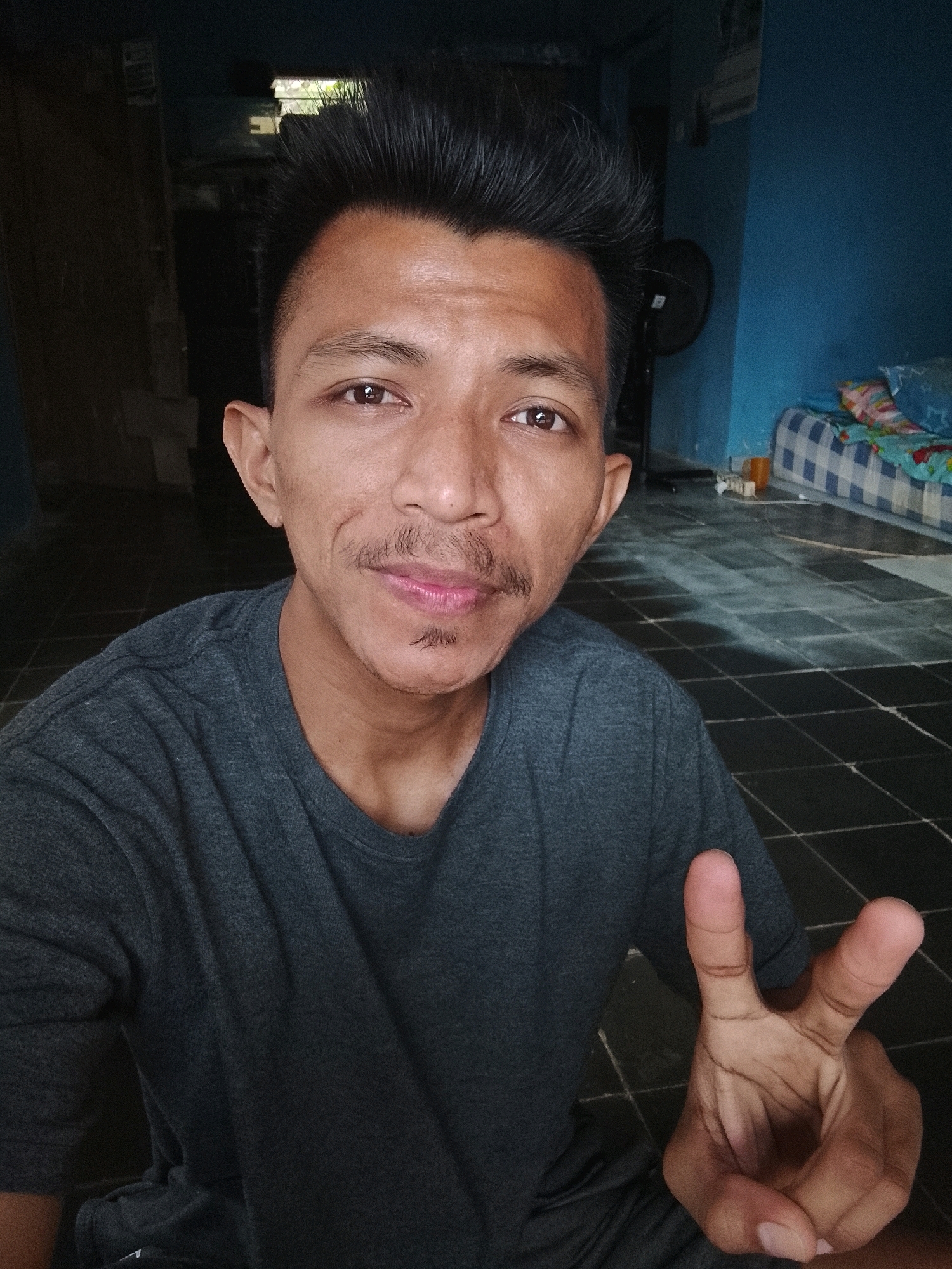 A young man is taking a selfie in an indoor environment. He is wearing a dark grey t-shirt and has short, dark, spiky hair. His right hand is raised, forming a peace sign. He has a thin mustache and a small beard. The background includes a blue wall, a bed with checkered bedding, and a black tiled floor. The lighting suggests it is daytime, with sunlight casting shadows. The location is identified as Kertahayu, Indonesia.