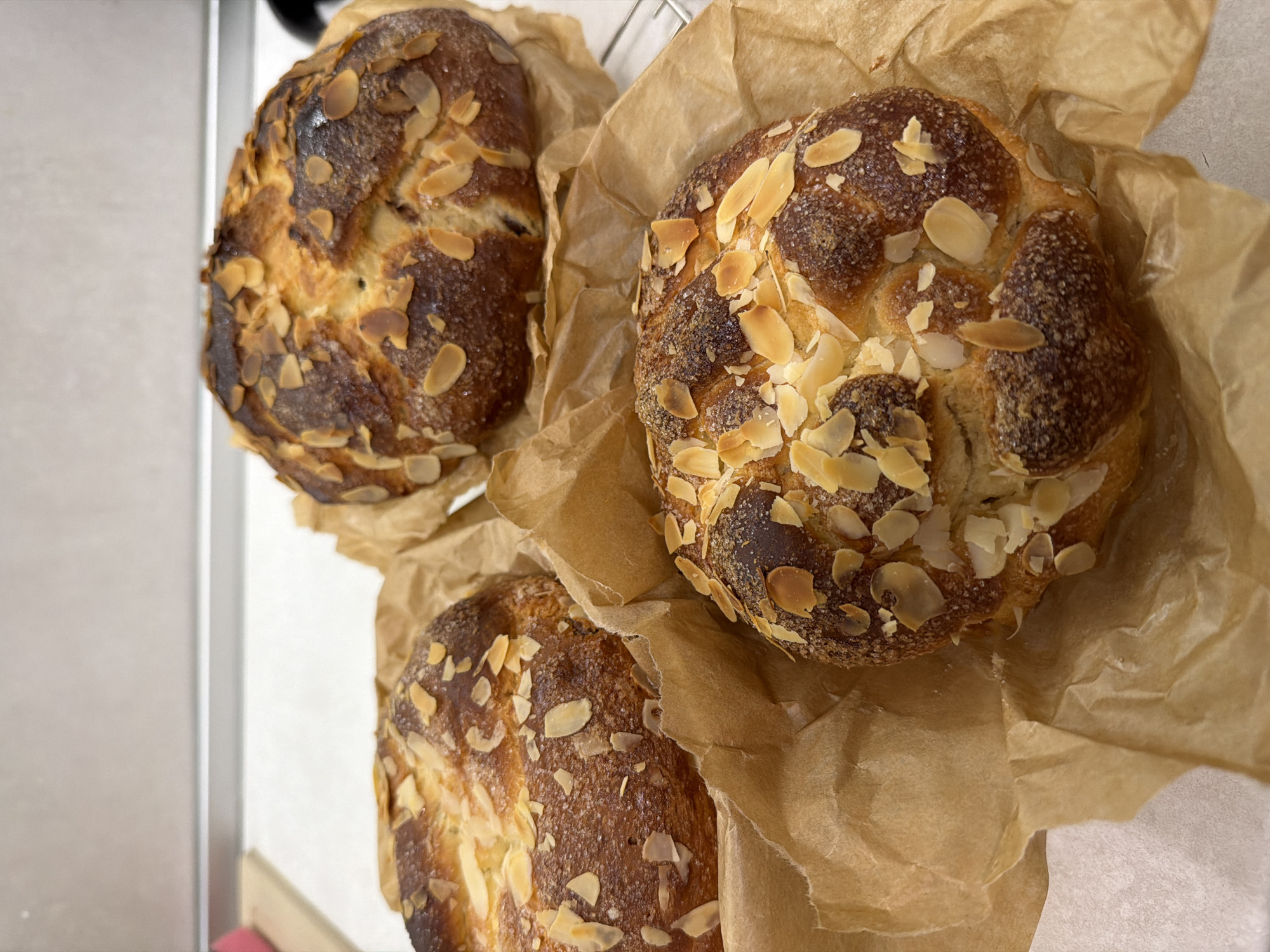 Three loaves of bread are arranged on a surface. The loaf on the right is nestled in crumpled brown paper. All three loaves are decorated with sliced almonds. The background shows a white surface, possibly a countertop or table, and a partial view of a white refrigerator or cabinet. This scene does not appear to be in Plovdiv, Bulgaria, as there are no elements in the image indicating that specific location.