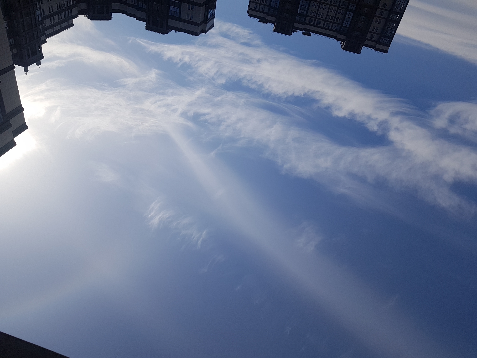 The image shows a sky view, possibly in Saint Petersburg, Russia, with buildings appearing at the top of the frame. The sky is blue, with thin, wispy clouds stretched across the field of view. The architecture of the buildings suggests a city location. No people are visible. The angle suggests the picture was taken looking upwards.