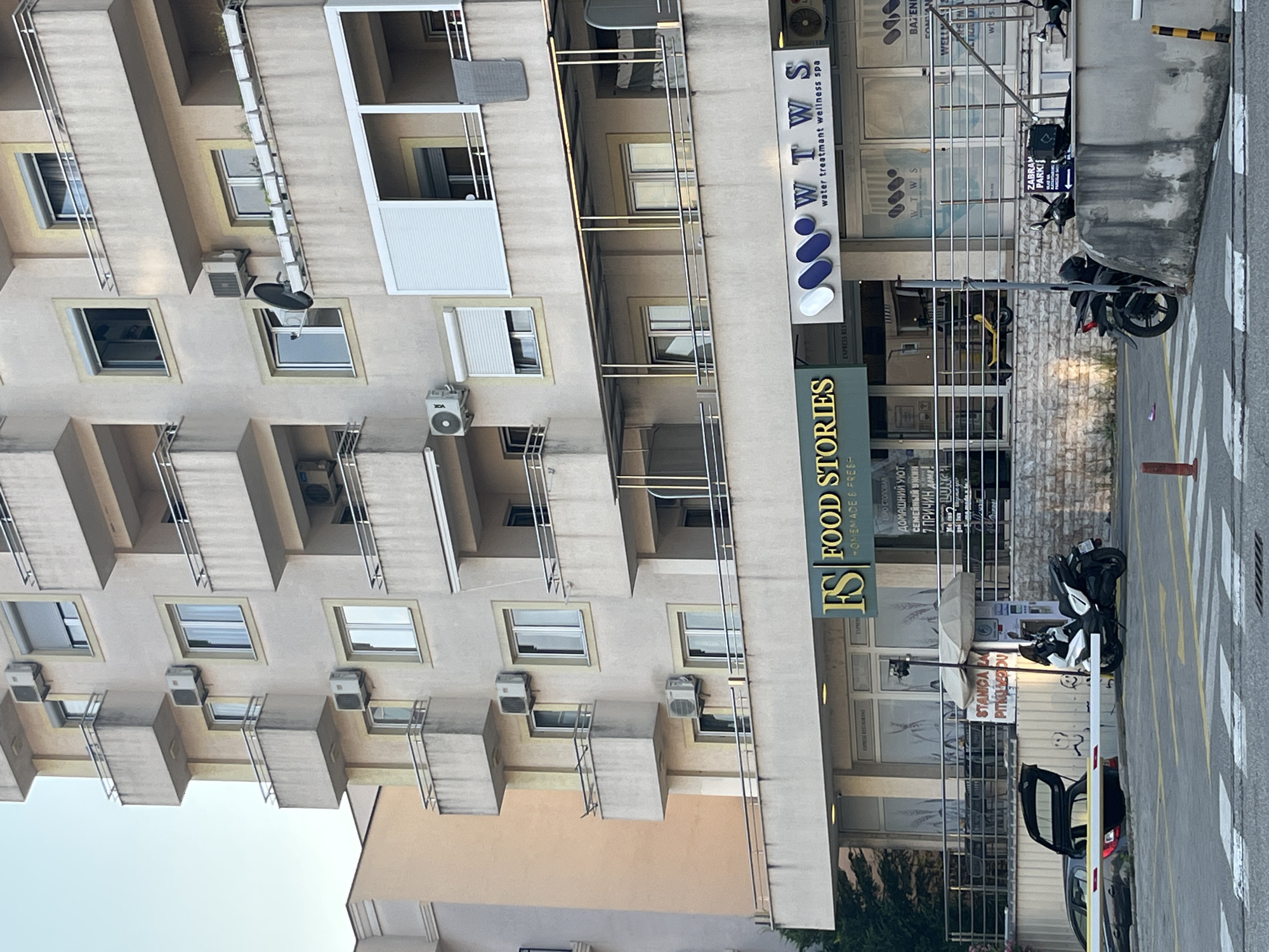 The image shows a narrow street scene in Budva, Montenegro, with a multi-story building on the left and a commercial establishment on the right. The building features a repetitive design of balconies with metal railings and windows, with several air conditioning units visible on the exterior walls. The commercial establishment on the right has a storefront with a large glass facade, displaying signs for "FS FOOD STORIES HOMEMADE & FRESH" and "WTWS water treatmant wellness spa." Other text in Cyrillic script is also visible, suggesting a Slavic language presence.
The street itself appears to be a pedestrian walkway or a service area, with yellow and white striped markings indicating a crosswalk or parking bay. Several motorcycles are parked near the storefront, and a car with its trunk open is visible in the foreground, partially behind a barrier. The lighting suggests it is daytime, with diffused sunlight casting soft shadows. The overall impression is that of a bustling urban environment, likely a commercial district, with a mix of residential and service-oriented businesses. The presence of multiple signs in different languages indicates a tourist-friendly location. There are no people prominently featured in the image.