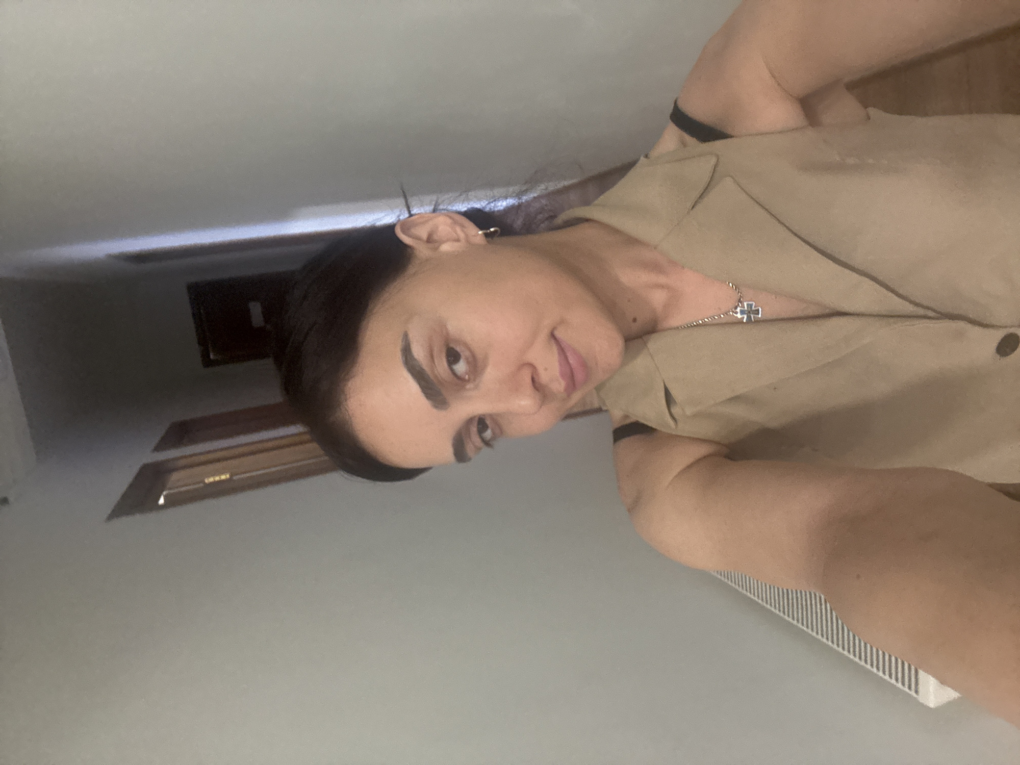 A selfie shows a woman in a beige vest, wearing a silver cross necklace. She is standing in a hallway with a radiator on the left side of the frame. In the background, there are two doorways. The image is taken from a slightly upward angle.