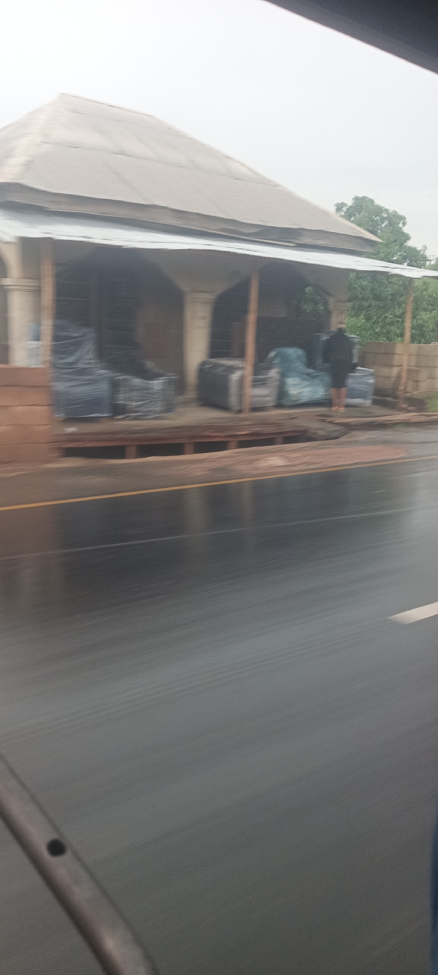 The photo depicts a furniture store in Akoli Imenyi, Nigeria, located along a street. The building has a gable roof and an open storefront supported by columns. Inside the store, furniture items are visible, some wrapped in plastic. A person in dark clothing stands in front of the store near concrete blocks. The road in front of the store is wet, with a visible yellow line dividing lanes. Motion blur suggests the photo was taken from a moving vehicle.