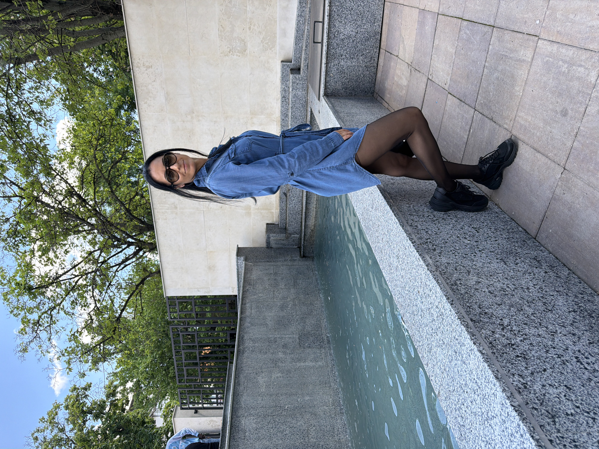 A woman with long black hair and sunglasses is reclining at the edge of a fountain in what appears to be an urban setting. She is wearing a blue denim dress-shirt, black tights, and black sneakers. The fountain's rim is made of gray stone, and the water is visible. The background includes a light-colored stone wall and green trees. Based on the architecture and appearance of the surroundings, this location is likely in Pleven, Bulgaria.