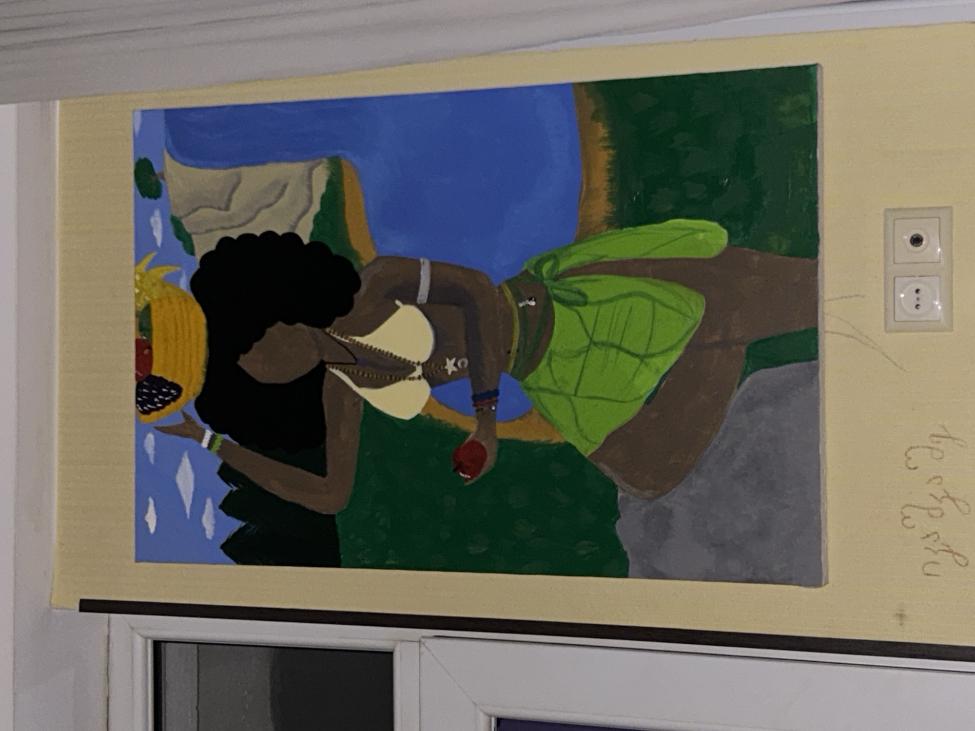 A painting of a woman with dark skin and a large afro hairstyle is depicted standing on a gray rock. She is wearing a yellow bikini top, a green wrap skirt, and jewelry. In her left hand, she holds a red apple, and on her head, she carries a basket filled with fruit, including grapes and strawberries. The background features a waterfall cascading into blue water, a green landscape with trees, and a bright blue sky with white clouds. The painting is framed with a light yellow border. Below the painting, there is writing in a script that appears to be Georgian, and a double electrical outlet is visible on the wall.