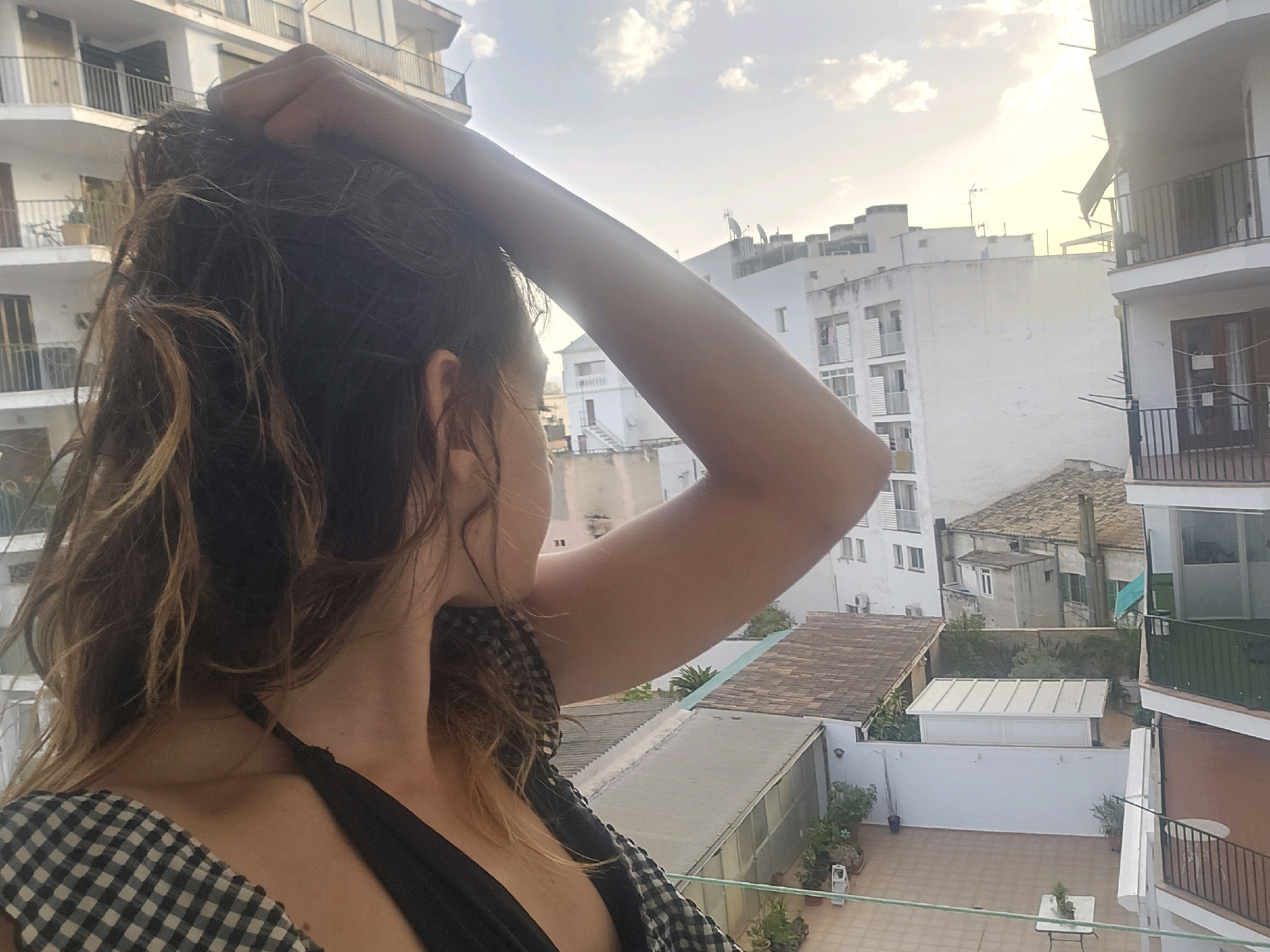 A woman, viewed from the shoulders up and over her right shoulder, stands on a balcony overlooking a courtyard in Palma de Mallorca, Spain. She is holding her right hand to the back of her head, her hair is partially up. She is wearing a black and white gingham-print top with a black strap visible. The environment is urban, with buildings of varying heights and styles visible in the background. The sky is bright with clouds. A courtyard with a tiled floor, potted plants, and a small white table is visible below.