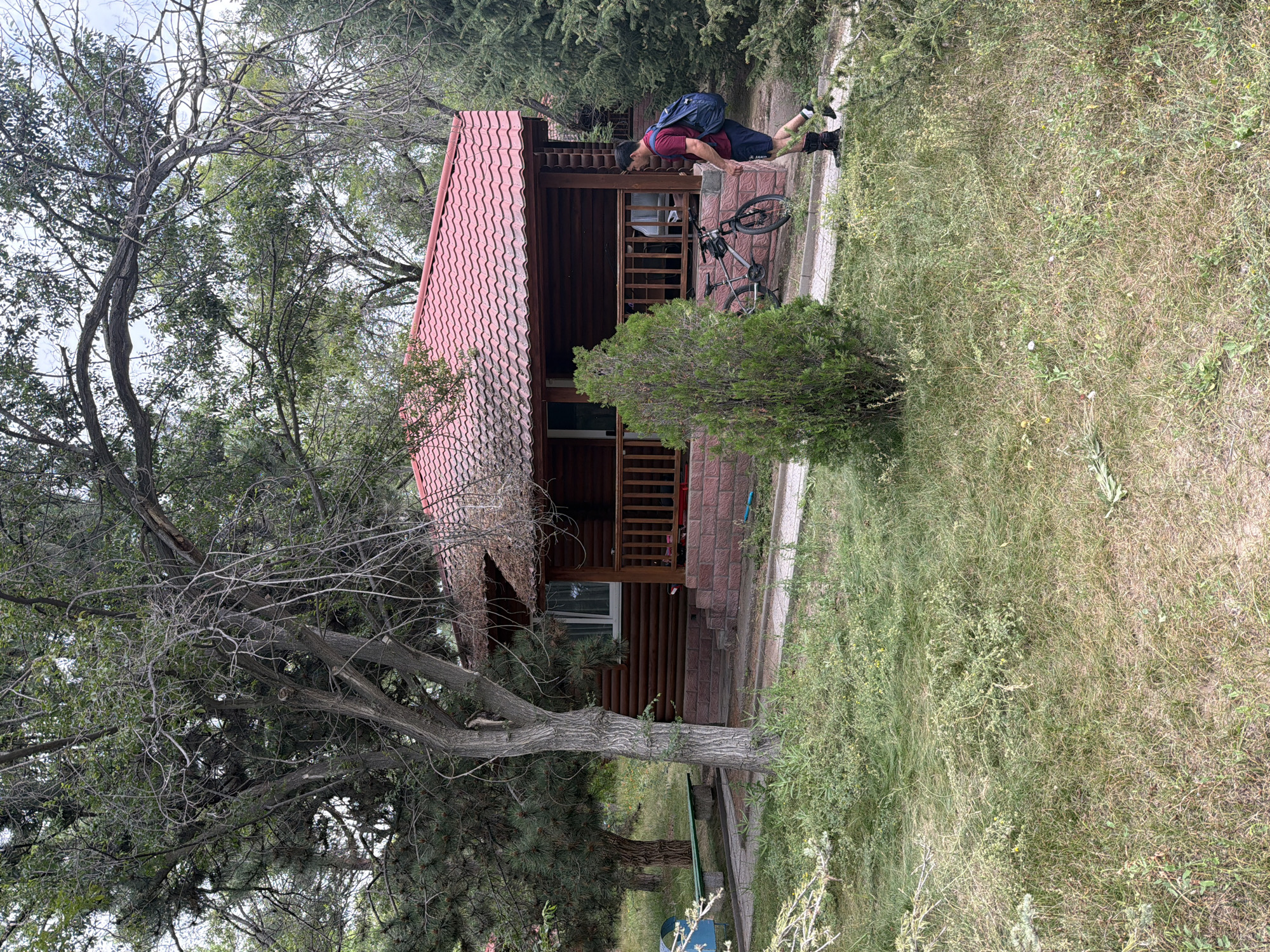 A man in a red t-shirt and blue shorts walks on a paved path next to a wooden cabin. The cabin has a red tiled roof and a wooden porch. A bicycle is parked next to the cabin. The area is surrounded by trees and grass. The man is carrying a blue backpack. The image is taken from a low angle.