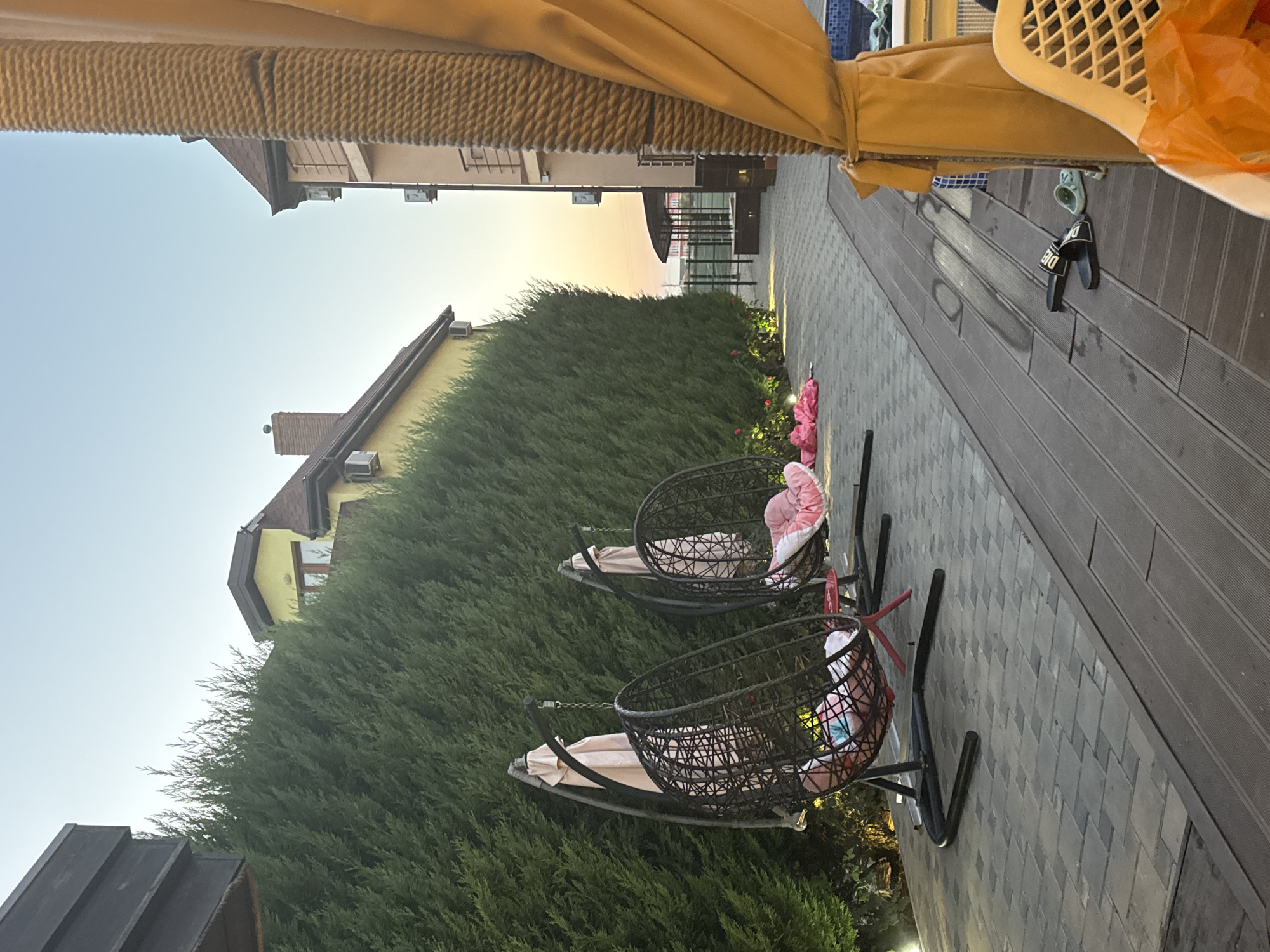 The image shows an outdoor patio area in Novofedorovka, Ukraine, in the late afternoon or early evening. Two hanging egg chairs, made of dark woven material with red and pink cushions, are positioned on a paved patio in front of a tall, dense green hedge. The hedge runs along the side of a yellow building with a brown roof and an additional tan building with balconies. A light-colored canopy or awning is visible on the right side of the frame, obscuring part of the tan building. On the wooden decking in front of the patio, a pair of black flip-flops and a light blue slide are present. A white plastic chair and an orange plastic bag are also visible in the foreground on the right. The sky is clear and shows the soft colors of sunset. The scene appears to be a private residence or resort.