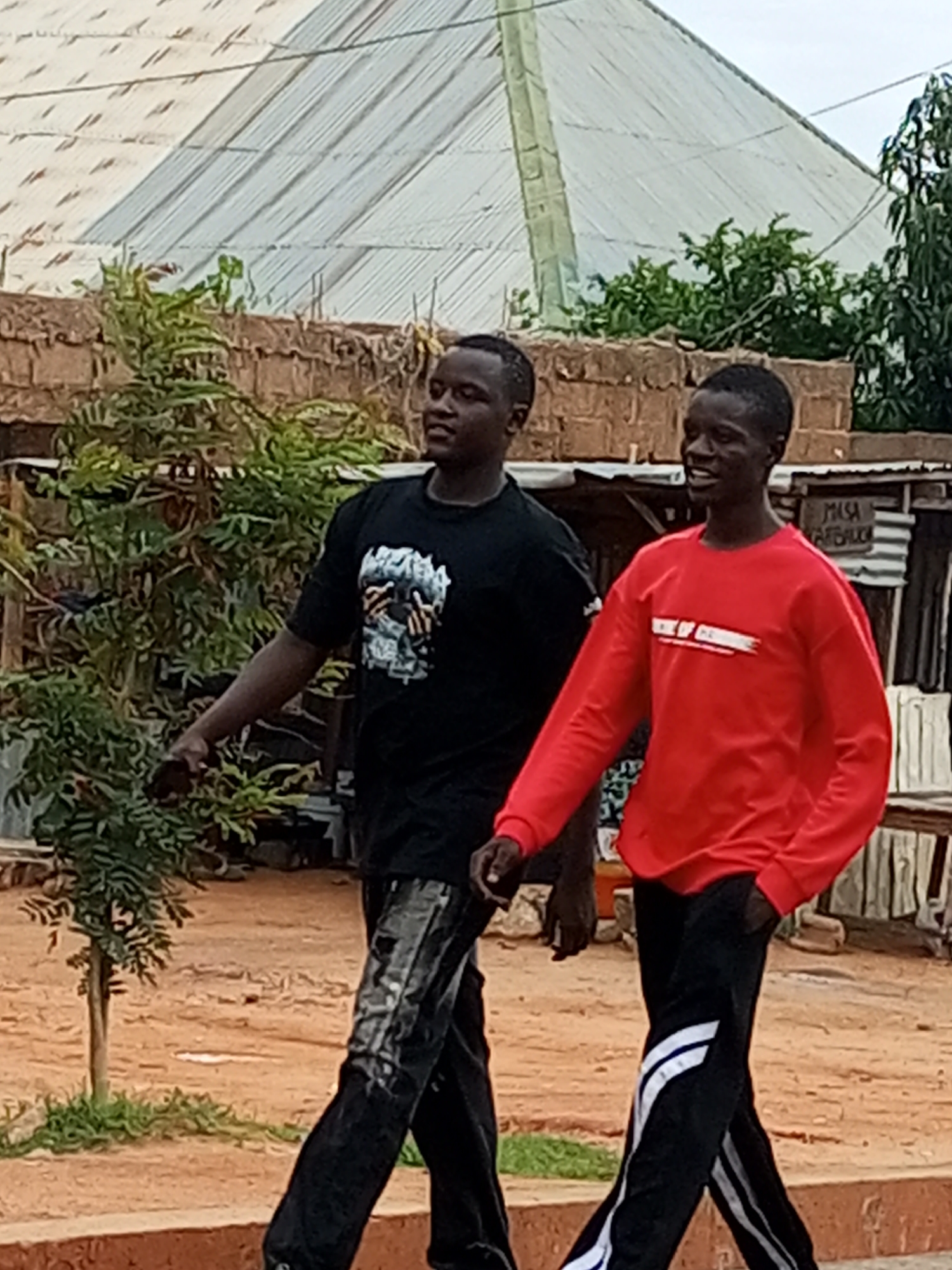 Two young men walk from left to right in the foreground of the image. The man on the left is wearing a black t-shirt with a graphic print and black pants with a shiny, patterned texture. The man on the right is wearing a red long-sleeved shirt with white text and black pants with white stripes. They are walking on a reddish-brown dirt path in front of a low brick wall and some vegetation. In the background, there is a large structure with a corrugated metal roof, appearing to be a greenhouse or market stall. The setting is outdoors in what appears to be a rural or semi-urban area in Tempure, Nigeria. The sky is overcast.