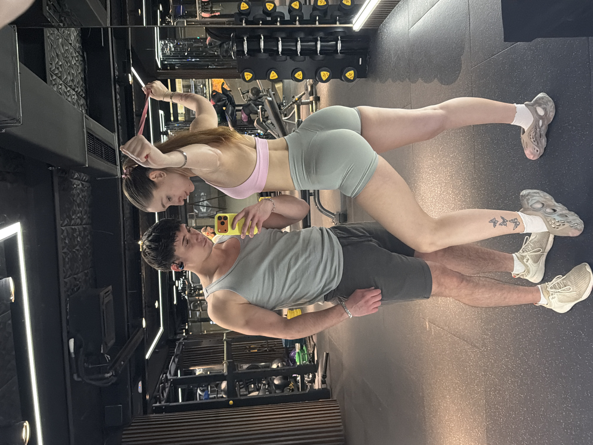 The image shows a man and woman in a gym, likely in Sofia, Bulgaria based on the architectural style sometimes seen in urban gyms there. They are posing for a selfie in front of a mirror. The woman is wearing a pink sports bra and light green shorts, and is holding a pink resistance band. The man is wearing a grey tank top and dark grey shorts, and is holding a yellow smartphone to take the photo. Gym equipment, including weights and machines, is visible in the background.