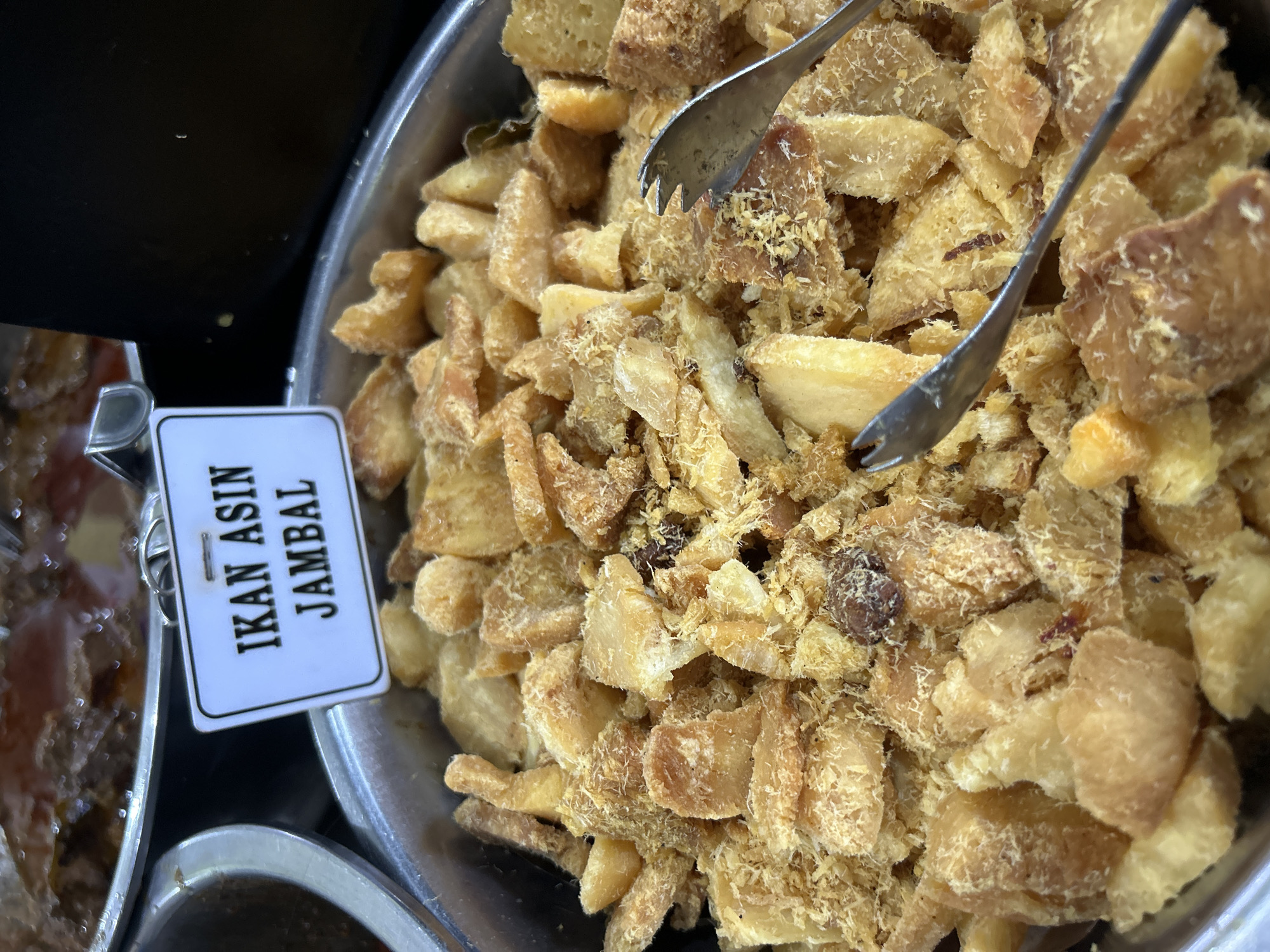The image shows a close-up view of a metal tray filled with fried fish, labeled "IKAN ASIN JAMBAL." The fish pieces are golden brown and appear to be a mixture of larger chunks and smaller, shredded pieces. Two metal serving tongs are visible in the tray, with one resting on top of the fish and the other partially submerged. In the background, out of focus, are other food containers, suggesting a buffet or food stall setting. The sign indicates that the food is likely found in Cirebon, Indonesia, a city known for its culinary traditions, particularly its seafood dishes like "ikan asin."