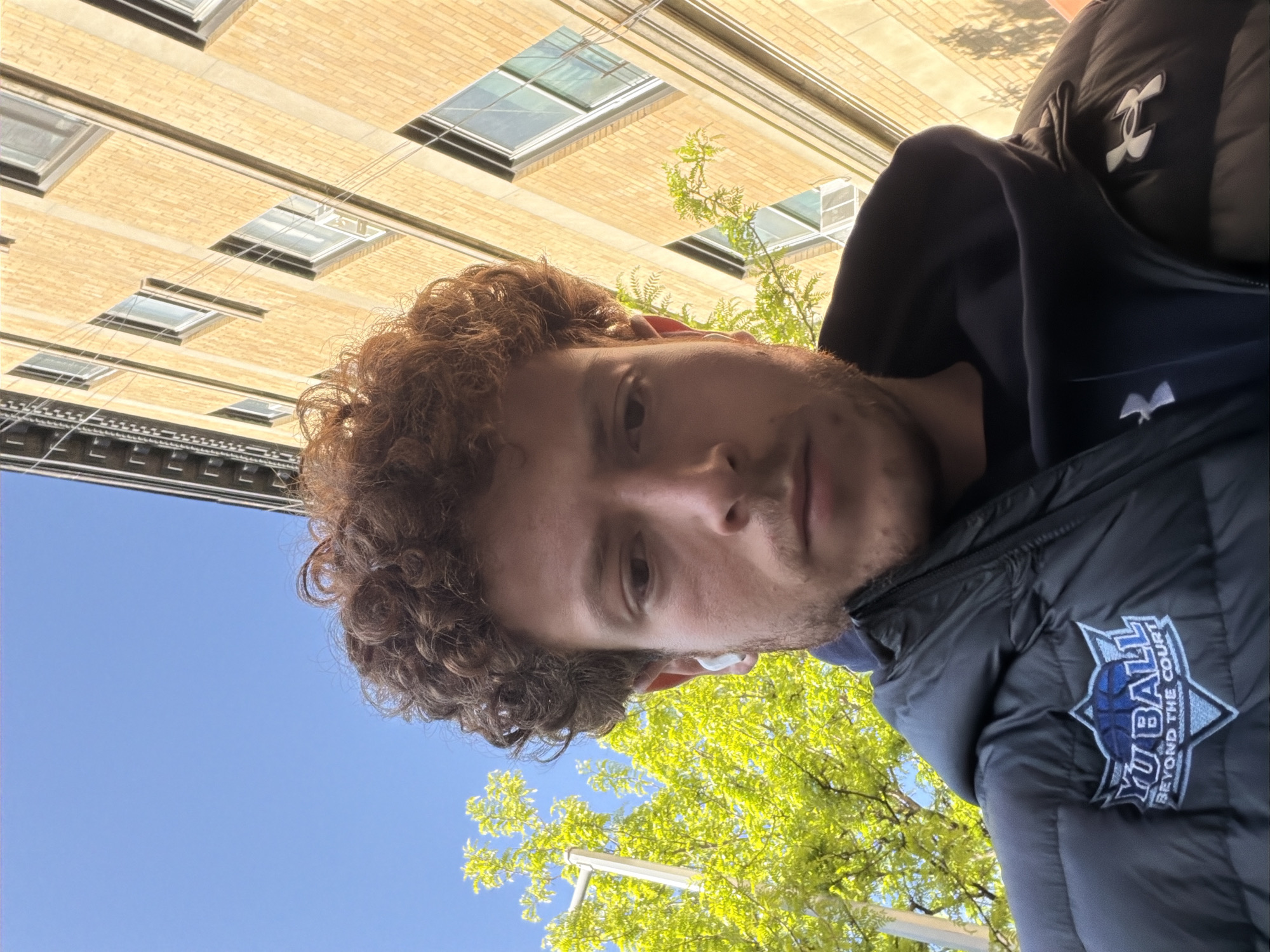 A portrait of a light-skinned male, with light brown curly hair, fair skin, and light stubble, looking directly at the camera, with the sky, trees, and a brick building as the background. The building has skylights and windows on it. The male is wearing a black jacket that has a logo on the right side of the chest. The logo has a basketball with “YU BALL BEYOND THE COURT” around it. The photo appears to be taken near Yeshiva University in New York.