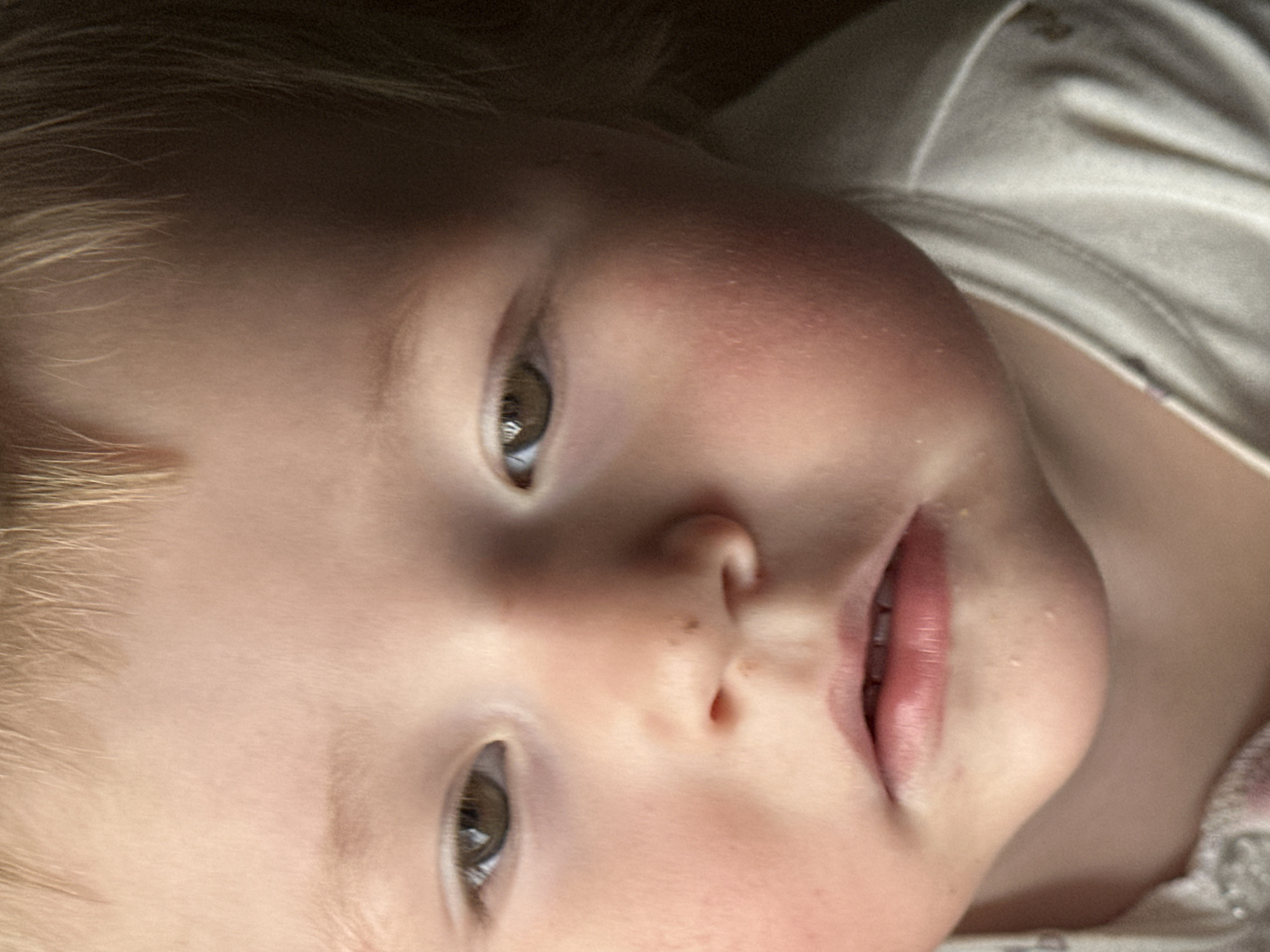 Close-up portrait of a young child with fair skin, light blonde hair, and hazel eyes. The child is lying on their back, facing the camera at an angle. The child is wearing a white top. The background is dark and out of focus. The scene does not appear to be in Nizhny Novgorod, Russia.