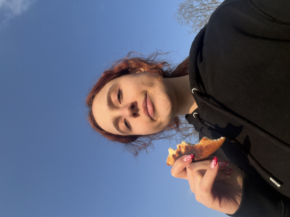 A young female, with reddish-brown hair pulled back, is depicted outdoors in Domgermain, France, on a clear day with a bright blue sky. She wears a black hooded sweatshirt with visible silver eyelets for the drawstrings. Her left hand, featuring manicured nails with a white base and red wavy designs, holds a partially consumed piece of golden-brown flatbread with a slightly charred crust. She is looking directly at the camera with a neutral to slightly amused expression. In the background, to the right, bare tree branches are visible against the sky. The lighting suggests natural sunlight from the left, casting subtle shadows on her face.
