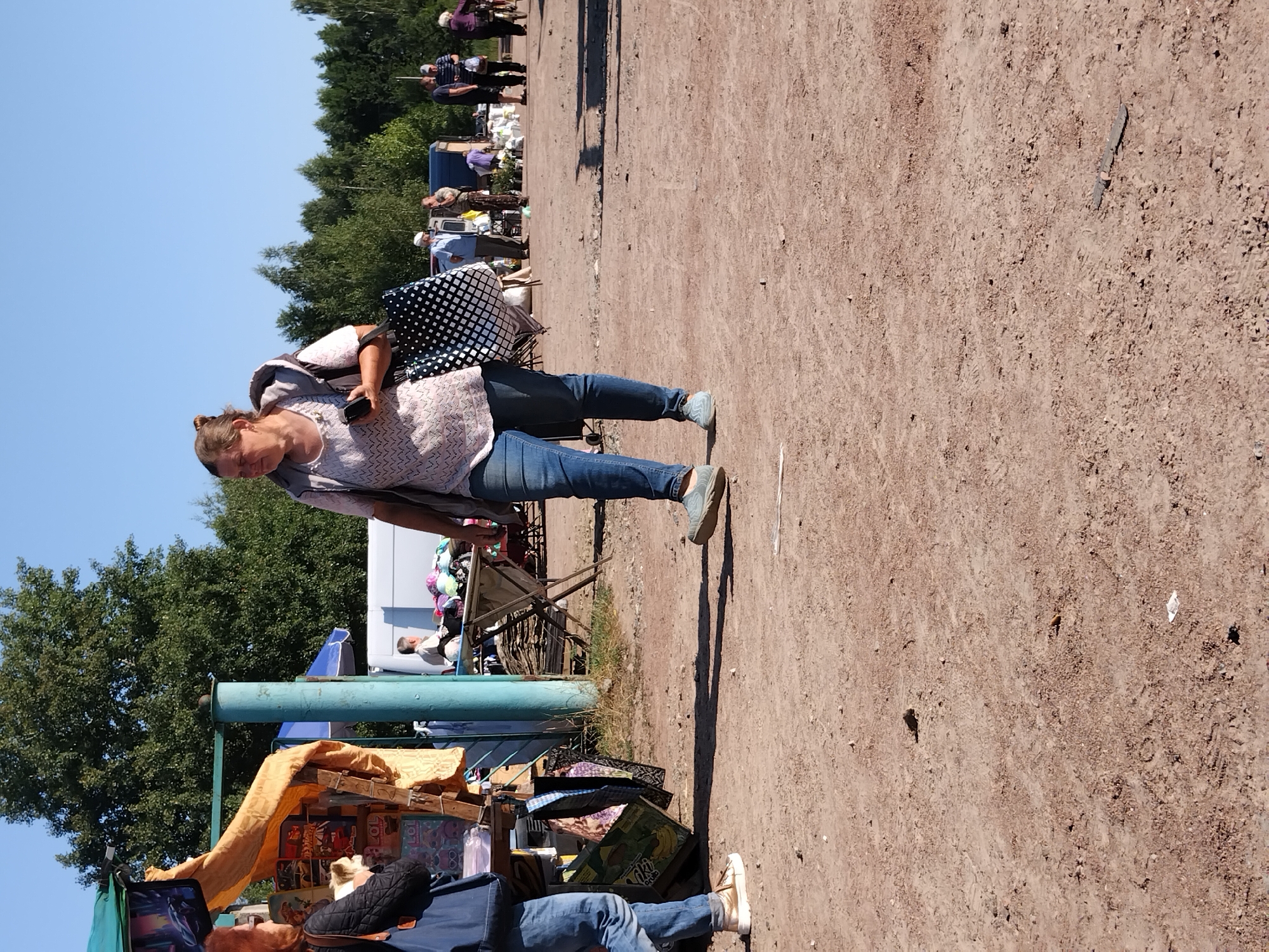 A woman walks across a dirt lot in a city, possibly Irshansk, Ukraine, carrying a shopping bag. She is wearing a light-colored, patterned top and blue jeans. Behind her, other people are browsing stalls at what appears to be an outdoor market. Trees line the background under a clear blue sky. The ground is mostly dirt with some patches of dry grass.