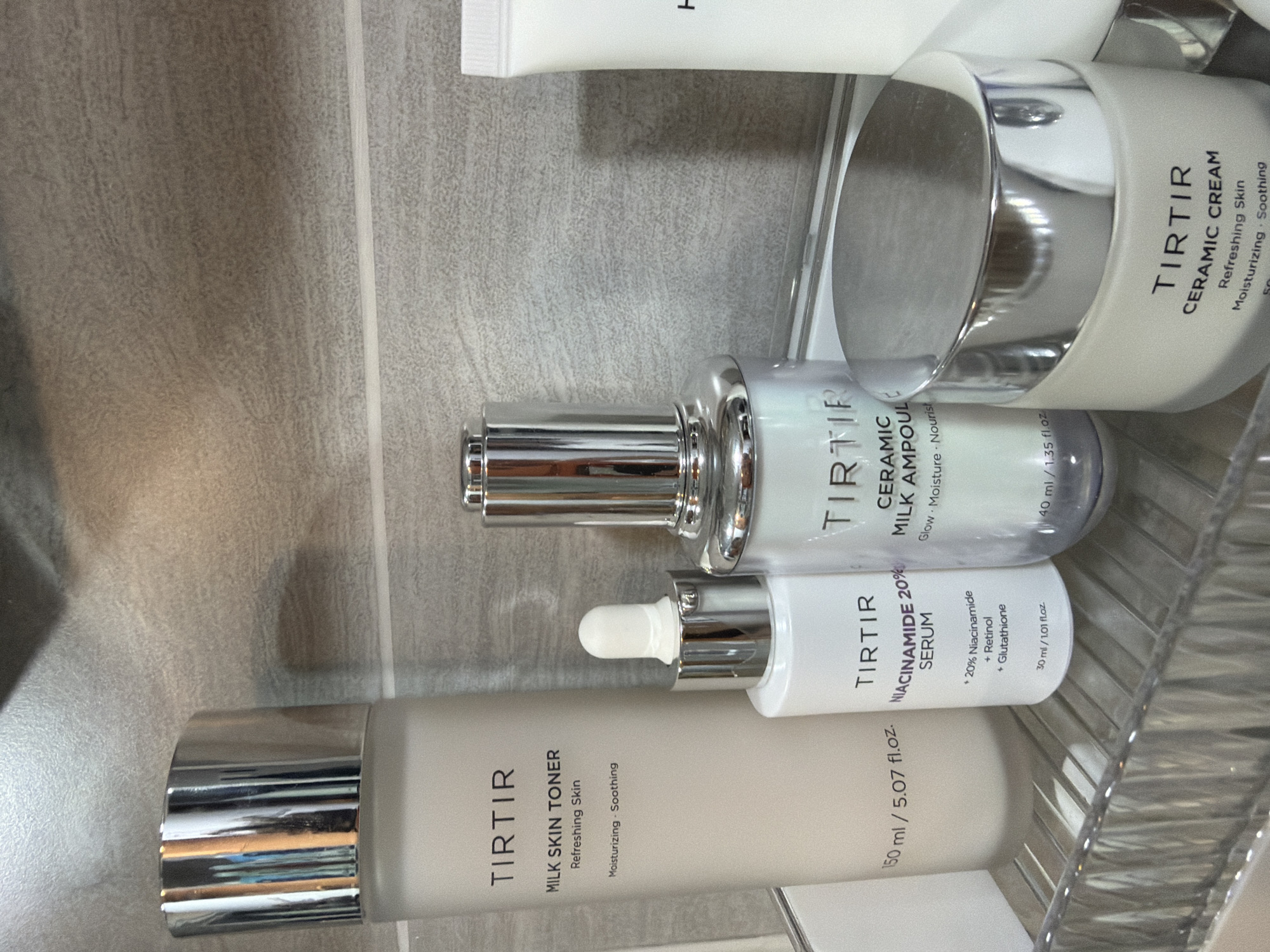 This image captures a static arrangement of five skincare products on a clear, ribbed plastic shelf, set against a light-colored, textured tiled wall, possibly in a bathroom environment. No individuals are present, nor are any actions depicted. The specific location in the city of Bangkok, Thailand, or any other geographical location, cannot be determined from the visual information provided.
The primary subjects are four visible products from the brand "TIRTIR," with a fifth product partially visible on the far right. From left to right:
1. A tall, cylindrical, frosted off-white bottle with a shiny silver, vertically ribbed cap, identified as "TIRTIR MILK SKIN TONER," stating "Refreshing Skin" and "Moisturizing - Soothing." Its volume is marked as "150 ml / 5.07 fl.oz."
2. A shorter, clear glass bottle with a silver collar and a white dropper bulb, labeled "TIRTIR NIACINAMIDE 20% SERUM." Subtext indicates key ingredients as "+ 20% Niacinamide + Retinol + Glutathione," with a volume of "30 ml / 1.01 fl.oz."
3. A medium-height, clear glass bottle with a shiny silver pump dispenser, partially visible text identifies it as "TIRTIR CERAMIC MILK AMPOUL," with descriptions "Glow - Moisture - Nourish" and a volume of "40 ml / 1.35 fl.oz."
4. A short, wide, frosted off-white jar with a broad, flat, shiny silver lid, labeled "TIRTIR CERAMIC CREAM." Visible text indicates "Refreshing Skin" and "Moisturizing - Soothing," with a partially visible volume of "50 ml."
5. A white tube, primarily obscured, is visible on the far right, with no discernible branding or product information.
The products are orderly arranged on a transparent, horizontally ribbed plastic organizer shelf. The background consists of light gray or beige textured tiles, possibly ceramic or stone, with a horizontal grout line positioned roughly in the middle of the frame. Artificial lighting creates reflections on the metallic caps and lids, and subtle shadows are cast behind the products.