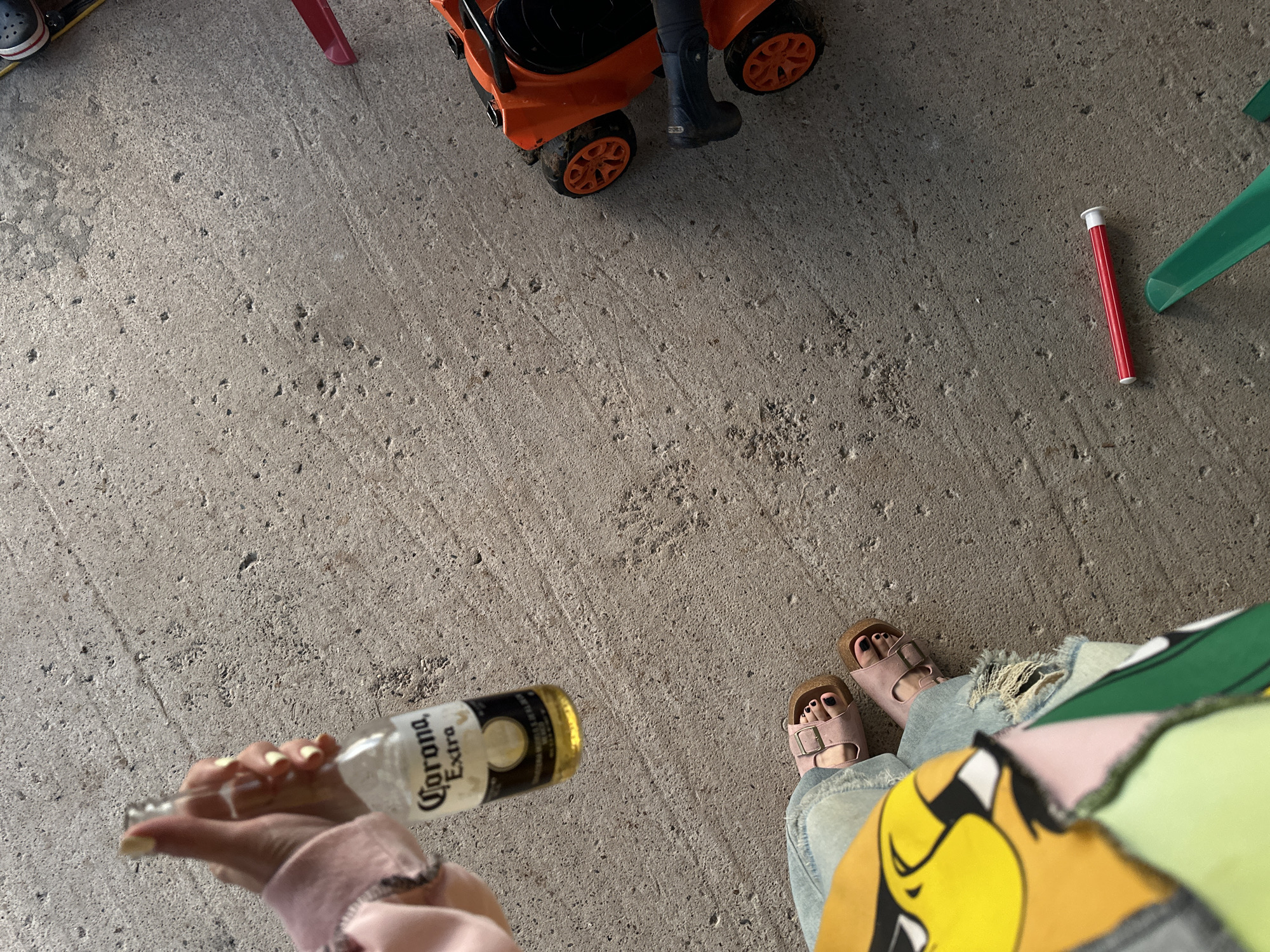The image captures a close-up, high-angle view of a concrete ground surface. In the upper left quadrant, a hand with pale skin and light yellow nail polish is holding a clear glass bottle of Corona Extra beer, with the label partially visible. Below this, a person's legs are visible, wearing light blue distressed jeans and pink Birkenstock sandals. The person is also wearing a brightly colored garment with a cartoonish yellow character, possibly part of a shirt or jacket. To the right of the frame, an orange toy lawnmower with black accents and orange wheels is present. A person's lower leg, clad in a dark boot, is positioned next to the toy. In the lower right quadrant, a red cylindrical object with white caps on both ends lies on the concrete. Several green plastic objects, possibly parts of a toy or a game, are also visible in the lower right corner. The overall scene suggests an informal outdoor setting, possibly a backyard or patio. There is no identifiable information to place this scene in Пепеляева, Russia.