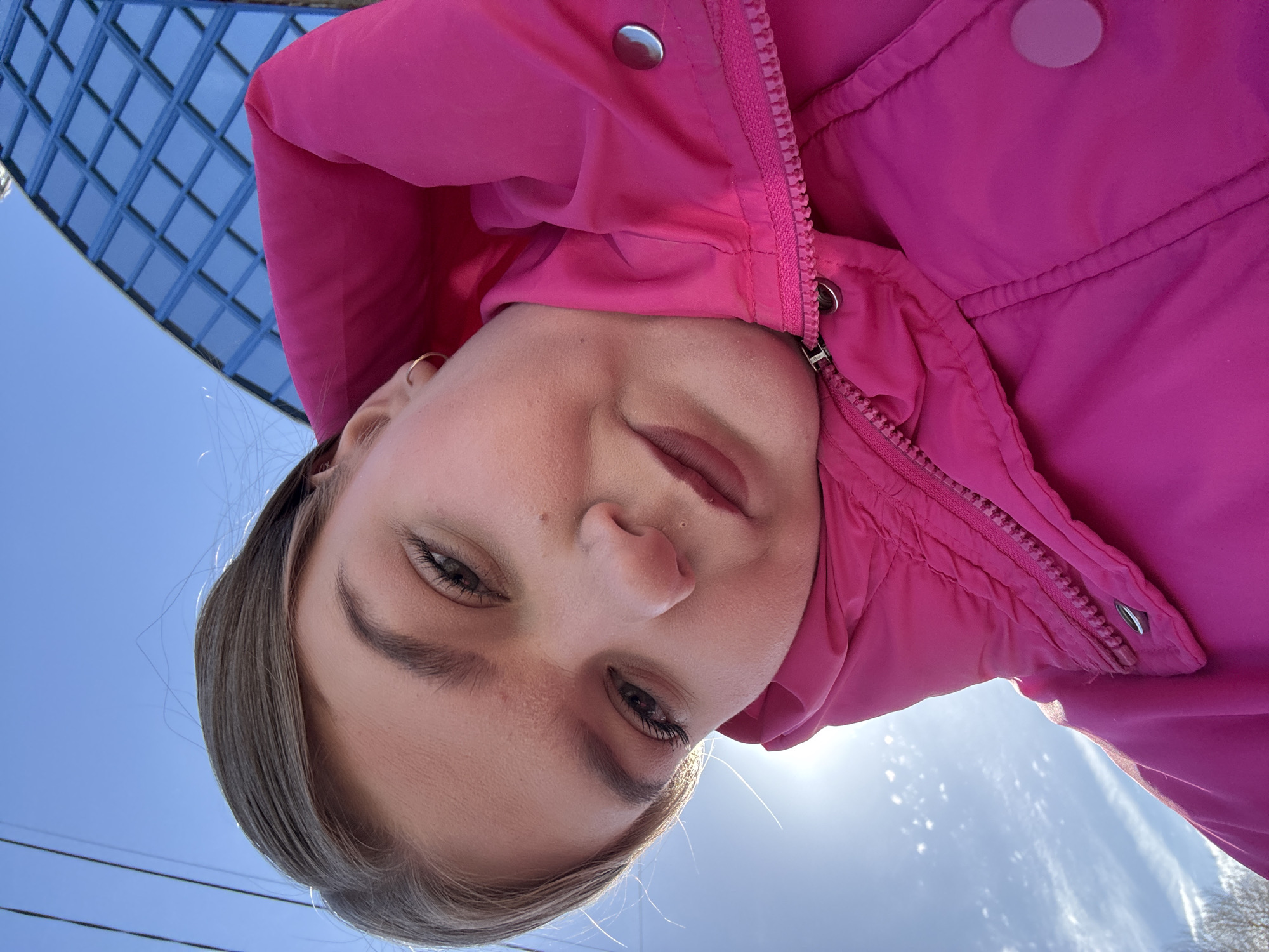 A close-up, low-angle selfie depicts a young female outdoors in Chaikovsky, Russia, captured on a clear, sunny day. The individual, positioned prominently in the frame from the chin upward, wears a vibrant pink padded jacket, partially zipped, featuring a high collar and a visible silver snap button on the lapel. She has light brown hair, mascara-defined eyelashes, groomed eyebrows, and dark red lipstick. A single gold hoop earring is visible in her right ear. Her expression is neutral, with a direct gaze toward the camera.
The background reveals a bright blue sky with sparse, wispy white clouds. The sun, a strong light source, creates a glare in the lower-left portion of the frame. Faint power lines or utility wires are visible against the sky on the far left. Dominating the upper-right background is a distinctive modern building with a blue, grid-patterned glass facade, identifiable as the Sports and Recreation Center "Ice Palace" (Ледовый дворец), located at ul. Karla Marksa, 27a, Chaykovsky, Perm Krai. Bare tree branches are partially visible in the extreme lower-left corner, beneath the sun's glare.