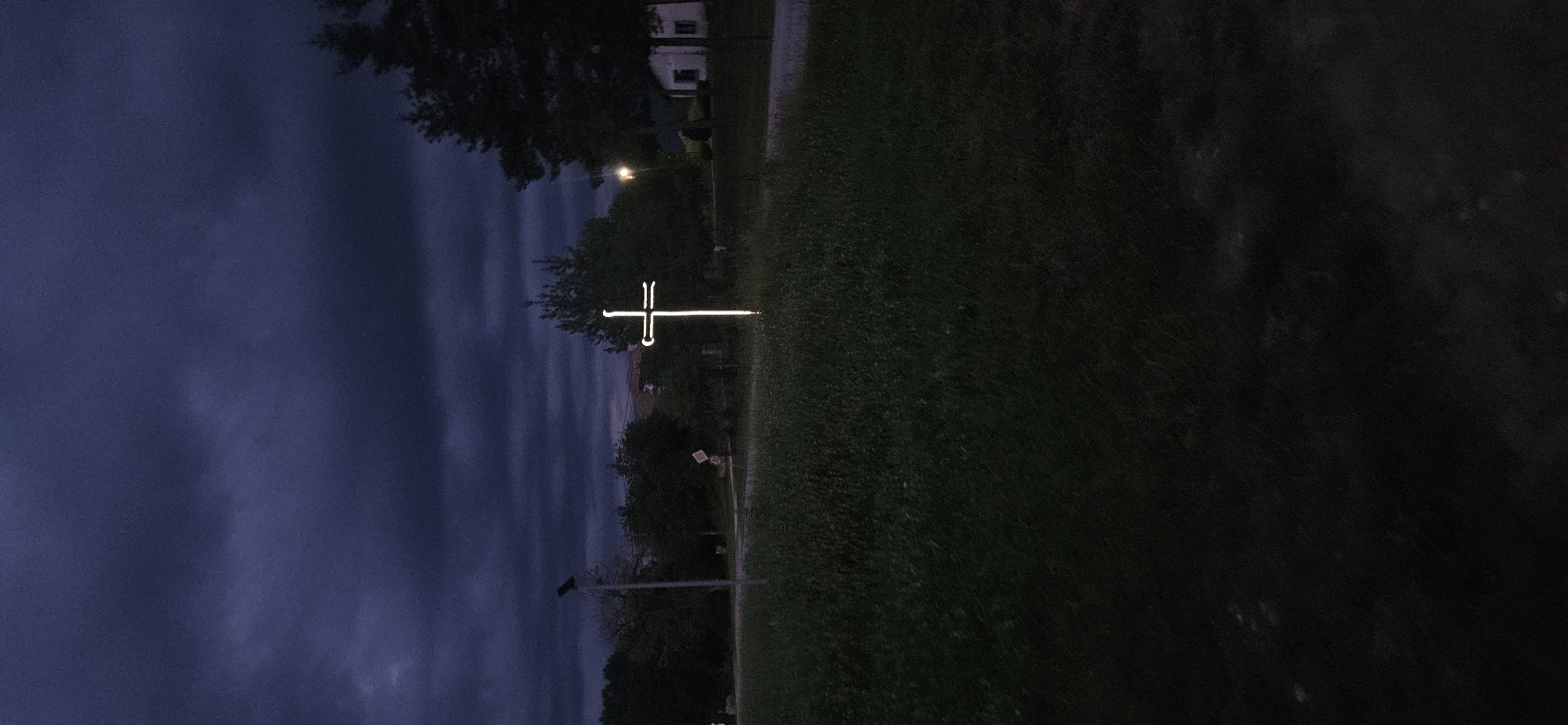 This image depicts an outdoor scene in Gutsal, Bulgaria, at dusk or night, characterized by low light and an overcast, dark blue-grey sky with heavy cloud cover. The central and most prominent object is a brightly illuminated, white, stylized Latin cross standing vertically in a field of dark green grass. The cross emits a strong, uniform light, with the horizontal arm featuring two distinct, short, inward-facing extensions or decorative elements near its ends.
To the left of the cross, a tall, dark grey utility pole with a solar panel on top stands amidst dark trees and vegetation. Further left, a circular white traffic sign with a red border and the number "30" in black indicates a speed limit. In the background, dark silhouettes of trees dominate the middle ground. Towards the right, a distant, dimly lit streetlight emits a warm yellow glow, and a small, light-colored building, possibly a house, is partially visible. Another dark tree stands closer to the right foreground. The foreground consists entirely of dark grass, showing minimal detail due to the low light. No people, vehicles, or explicit actions are visible within the frame.