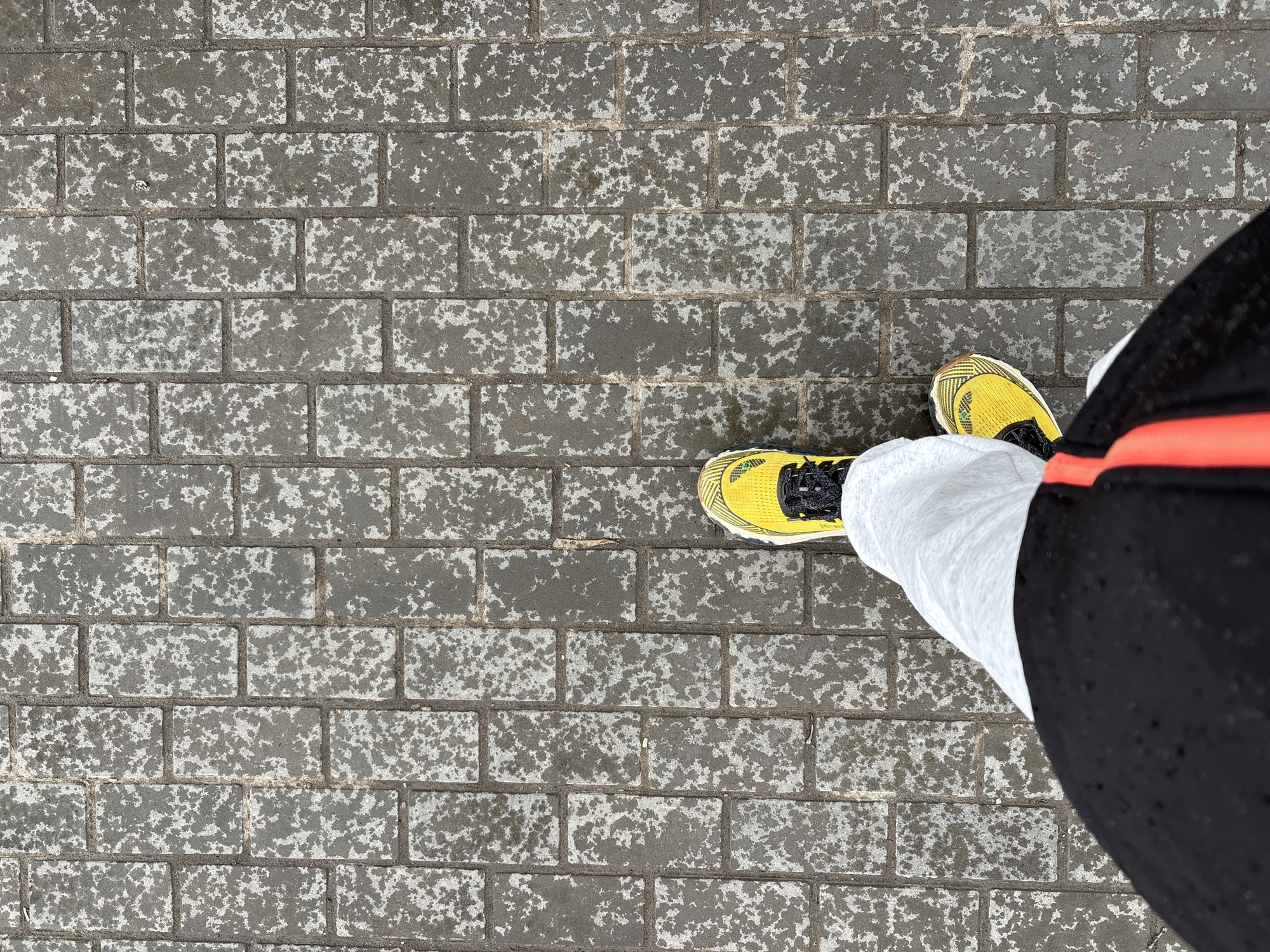 An overhead shot captures a person's lower legs and feet standing on a paved outdoor surface in Chaikovsky, Russia. The individual wears bright yellow running shoes with black accents and laces, paired with white socks. Part of their black shorts or pants, featuring a red side stripe, is also visible.
The ground is constructed from rectangular grey bricks or pavers laid in a stretcher bond pattern. A distinctive speckled white and light grey pattern covers the surface of these bricks, suggesting the presence of frost, light ice, or a wet, weathered texture. Dark grout lines separate the individual pavers.
The scene depicts the person standing still, with the lighting indicating it is daytime, likely under an overcast sky due to the absence of harsh shadows. The speckled ground pattern, combined with the location context, implies cool or cold weather conditions. No visible text or discernible emotions are present in the image.