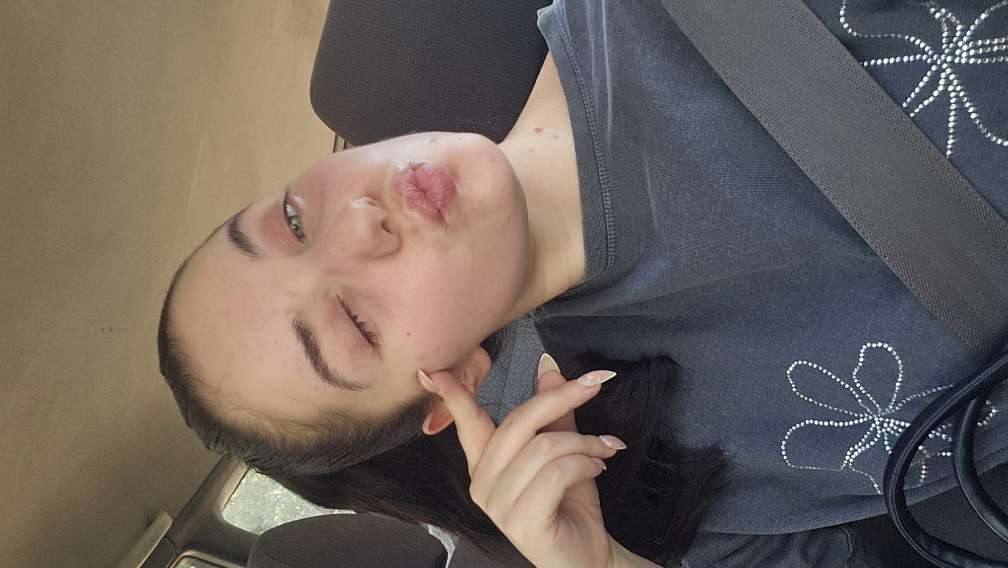A young adult female is depicted in a close-up portrait, seated inside a vehicle. She has dark hair pulled back into a ponytail, visible over her left shoulder. Her right eye is open, revealing a green or hazel iris, while her left eye is closed in a wink. Her lips are pursed into a kissy face. Her right hand is raised, with her index finger gently touching her right cheekbone. Her nails are long, pointed, and have a light-colored tip. She is wearing a dark gray, possibly faded, crew-neck t-shirt adorned with a sparkly, multi-petal flower design on the chest. A dark gray car seatbelt crosses her torso diagonally from her right shoulder. A segment of a dark bag or strap is visible in the lower-left corner. The car's interior features a light-colored fabric headliner and a window on the left, through which blurred green foliage is visible. The specific location in the city of Novakovo, Bulgaria, cannot be determined from the visual information provided in the image.