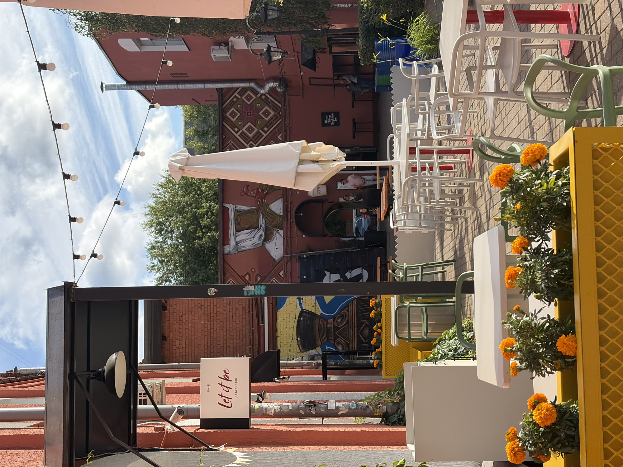 An outdoor seating area of a cafe named "Let it be" is depicted in this image. The cafe is located in Minsk, Belarus. Several tables and chairs, mostly white, are arranged on a brick-paved patio. Some tables have red bases, while others have white bases. In the foreground, a yellow planter box contains vibrant orange marigolds. Behind the tables, a white outdoor umbrella is partially folded. The background reveals a red building with a mural on its wall, depicting figures and geometric patterns. String lights are suspended across the open space overhead. A white sign with "Let it be" and Cyrillic text is visible on the left side of the image. Several people are casually scattered throughout the scene, some standing, some seated at tables. The sky is blue with scattered clouds.