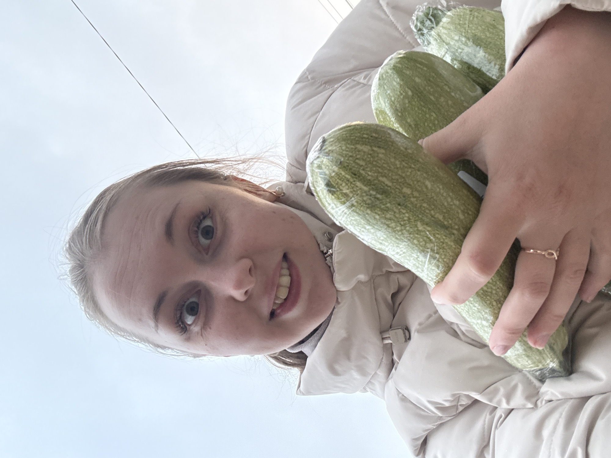 The image is presented in a portrait orientation, rotated 90 degrees clockwise. It captures a young woman from a low-angle perspective, looking down towards the camera with the sky as the primary background.
The woman has light-colored hair, likely pulled back, and her face occupies the left side of the frame. She is smiling broadly, revealing her teeth, and her eyes are open wide, looking directly at the viewer. Visible eyelashes suggest some eye makeup. She wears a light-colored, possibly beige or cream, padded or puffy jacket, with a grey collar or turtleneck visible underneath. A small, light-colored stud earring is present in her visible right ear (on the left side of the image).
In her right hand, which extends from the center towards the top right of the frame, she holds a bundle of at least three elongated, light green vegetables, resembling zucchinis or marrow squash. Each vegetable is individually wrapped in clear plastic film. A delicate, gold-toned ring with an open, infinity-like design is visible on her right ring finger.
The background is dominated by an expansive, bright light blue or grey sky, indicating an outdoor setting during daytime. A thin, dark line, possibly a power line or cable, stretches diagonally across the top right corner of the sky. The weather appears to be overcast or hazy. The woman's cheerful smile conveys a happy and content emotion. There is no visible text in the image.