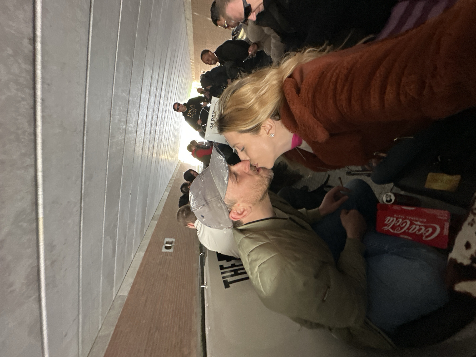 The image, taken in Amsterdam, shows a couple sharing an intimate kiss amidst a crowded indoor or covered outdoor space, resembling a corridor or waiting area. In the foreground, a man wearing a grey baseball cap, a green jacket, and blue jeans is seated, kissing a blonde woman in a brown fuzzy coat and a pink collar or scarf. The man rests one arm on his knee, while the woman leans in towards him.
Beside the man, a red Coca-Cola box and an open can are visible on the ground. The background is filled with other people, some seated, some standing, suggesting they are also waiting for an event or transportation. Many are looking forward or towards a focal point outside the frame. One man in the background wears a black hoodie with the text "DNA" and "44 PAY" visible. The surroundings feature light grey, vertically panelled concrete walls and a grey floor that transitions to a brown surface further back. Artificial lighting suggests an evening or nighttime setting. A partial sign in the foreground beneath the couple reads "THE" with subsequent letters obscured. The overall scene depicts a tender moment in a bustling public environment.