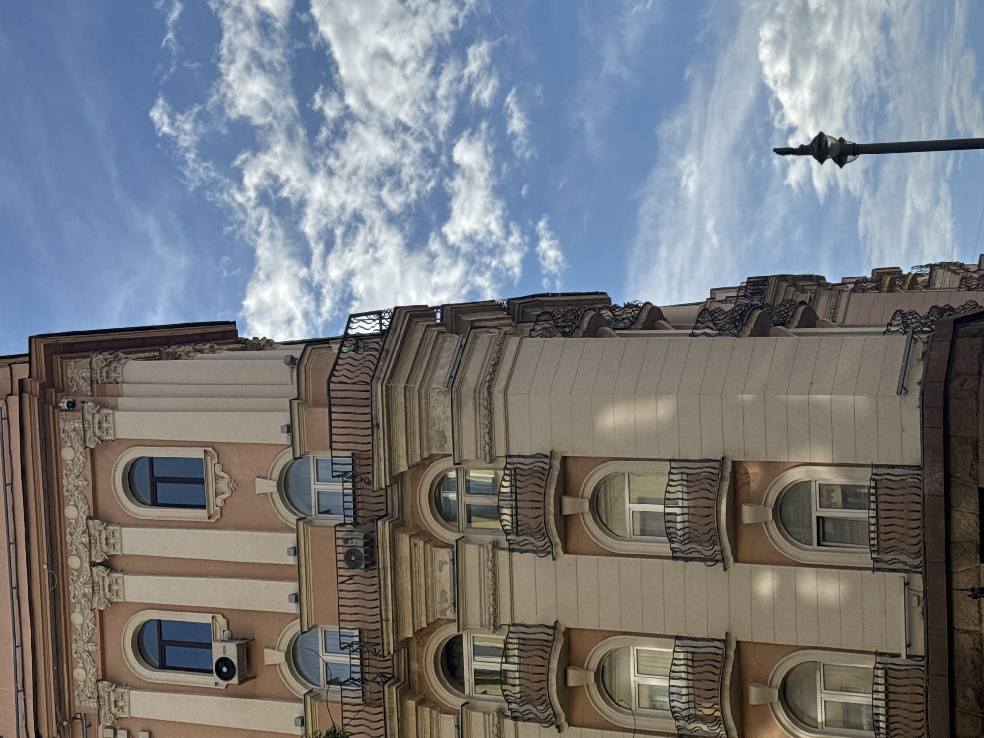 The image shows a building with ornate architectural details, including arched windows, balconies with decorative railings, and decorative molding. The building is beige with white trim, and it has an air conditioning unit installed in one of the windows. Above the building, there is a blue sky with scattered white clouds. A lamppost is visible in the upper right corner of the image. The building's style suggests it might be located in a European city, possibly Moscow, Russia, given its architecture.