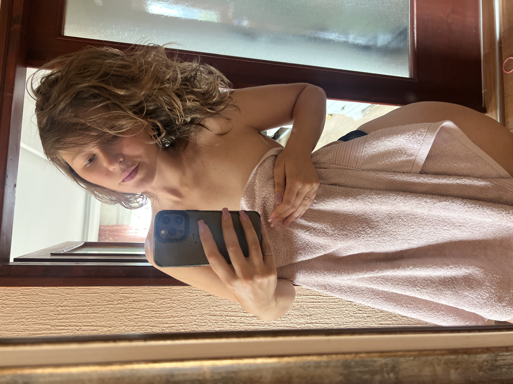 A young woman is taking a selfie in a mirror. She has light brown, wavy hair and is partially covered by a light pink towel. She is holding a smartphone with a green case. The mirror is framed by brown wood. The background suggests a room with a window. There is insufficient information in the image to determine the location in Podgorica, Montenegro.