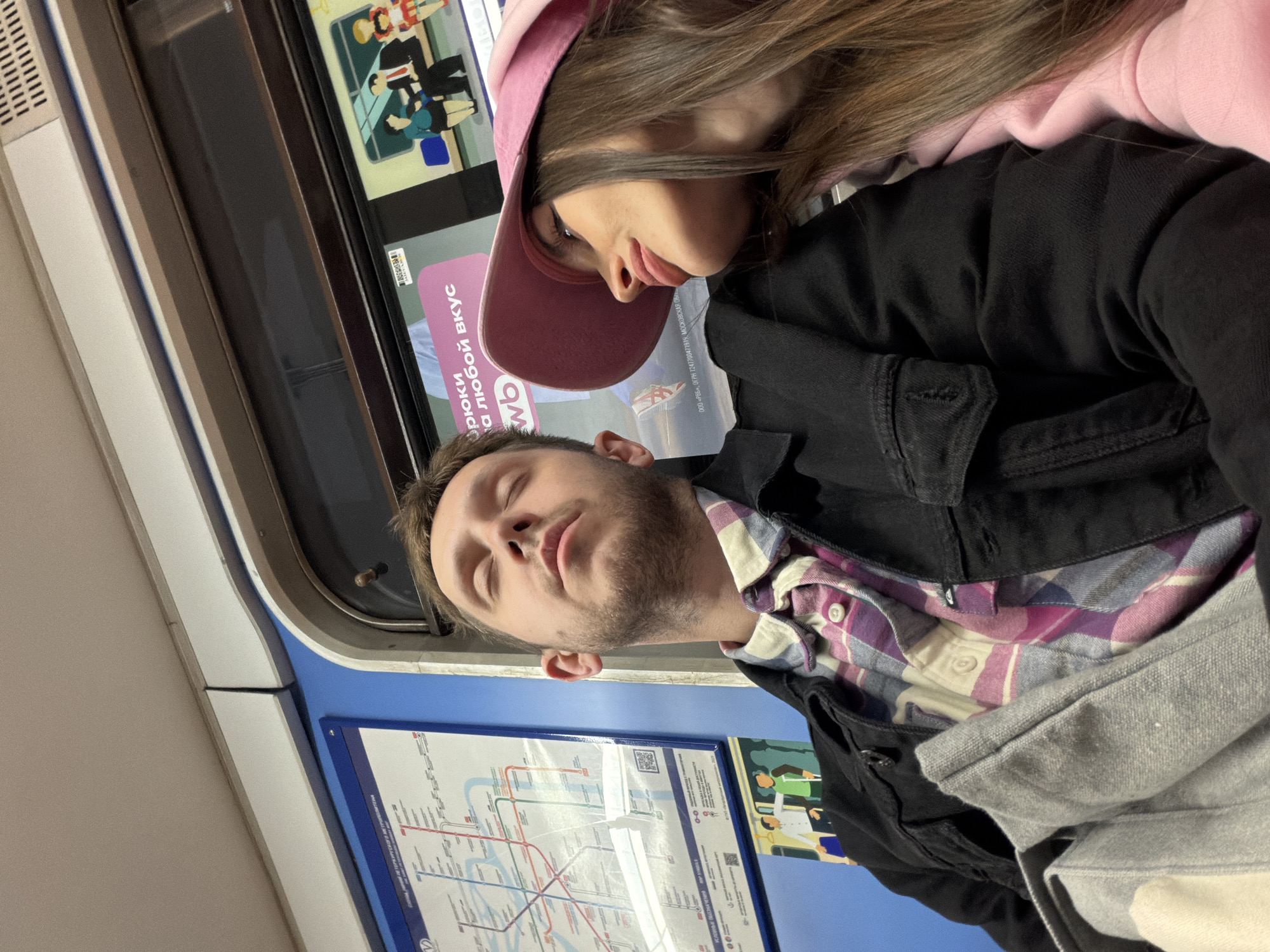 Here is a description of the image:
Inside what appears to be a subway car, a couple is seen posing for a picture. The man, with his eyes closed, has a plaid shirt and black jacket. A woman in a pink hat is closely positioned near him. Behind them is a window displaying an advertisement. A map of the subway system is also visible on the wall behind them. The map suggests the location is possibly Saint Petersburg, Russia.